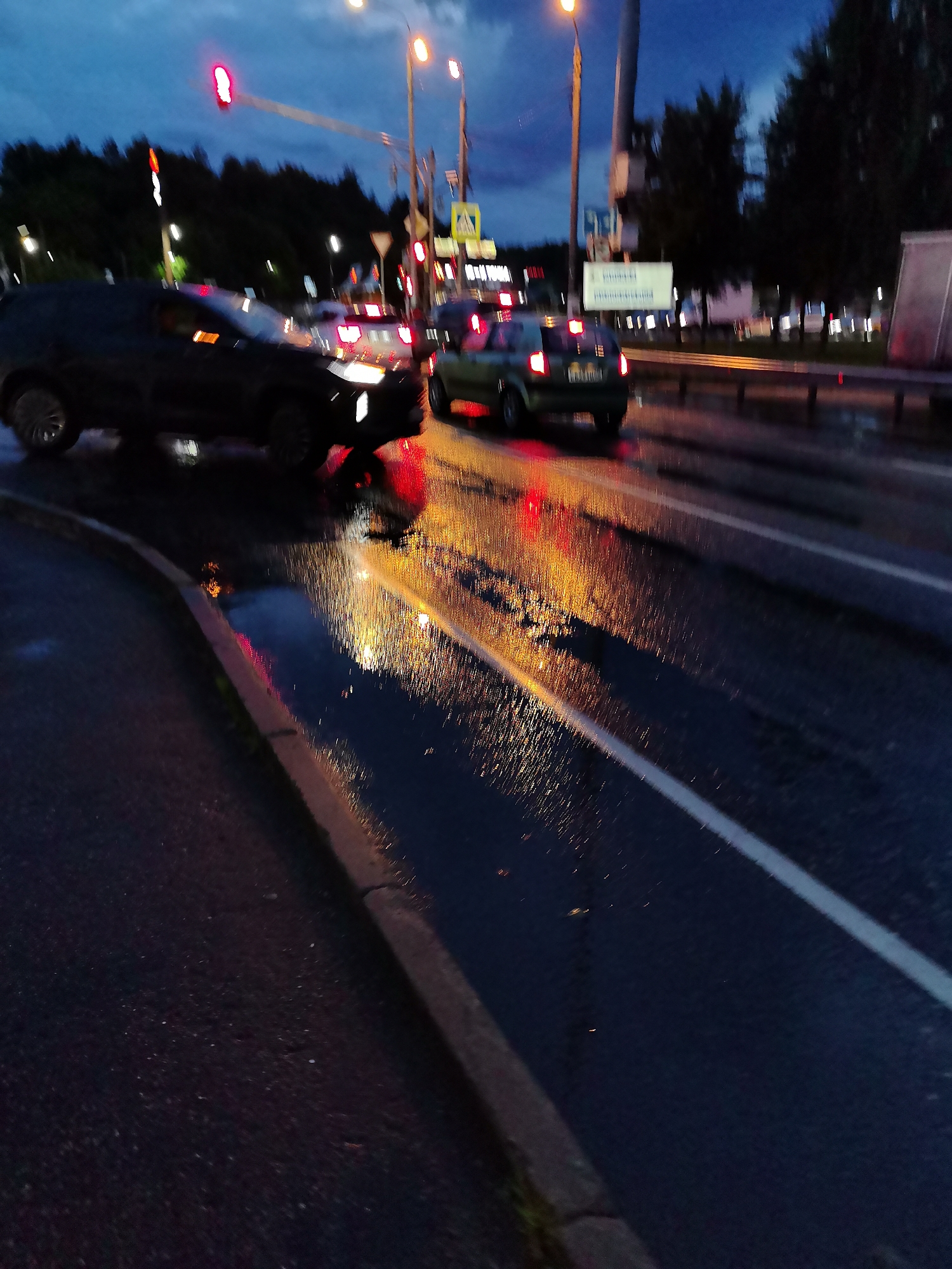 The image shows a dark, wet road at dusk or night, possibly in Krasnogorsk, Russia, with reflections of lights on the wet asphalt. A dark-colored SUV is positioned on the left side of the frame, appearing to be either stopped or moving slowly. Its headlights are on. To the right of the SUV, a lighter-colored car is visible in the lane of traffic, its brake lights illuminated. The road has clear white lane markings. Trees line the background, and traffic signals are visible overhead. The overall impression is of a street scene during evening or night with some traffic and wet conditions.