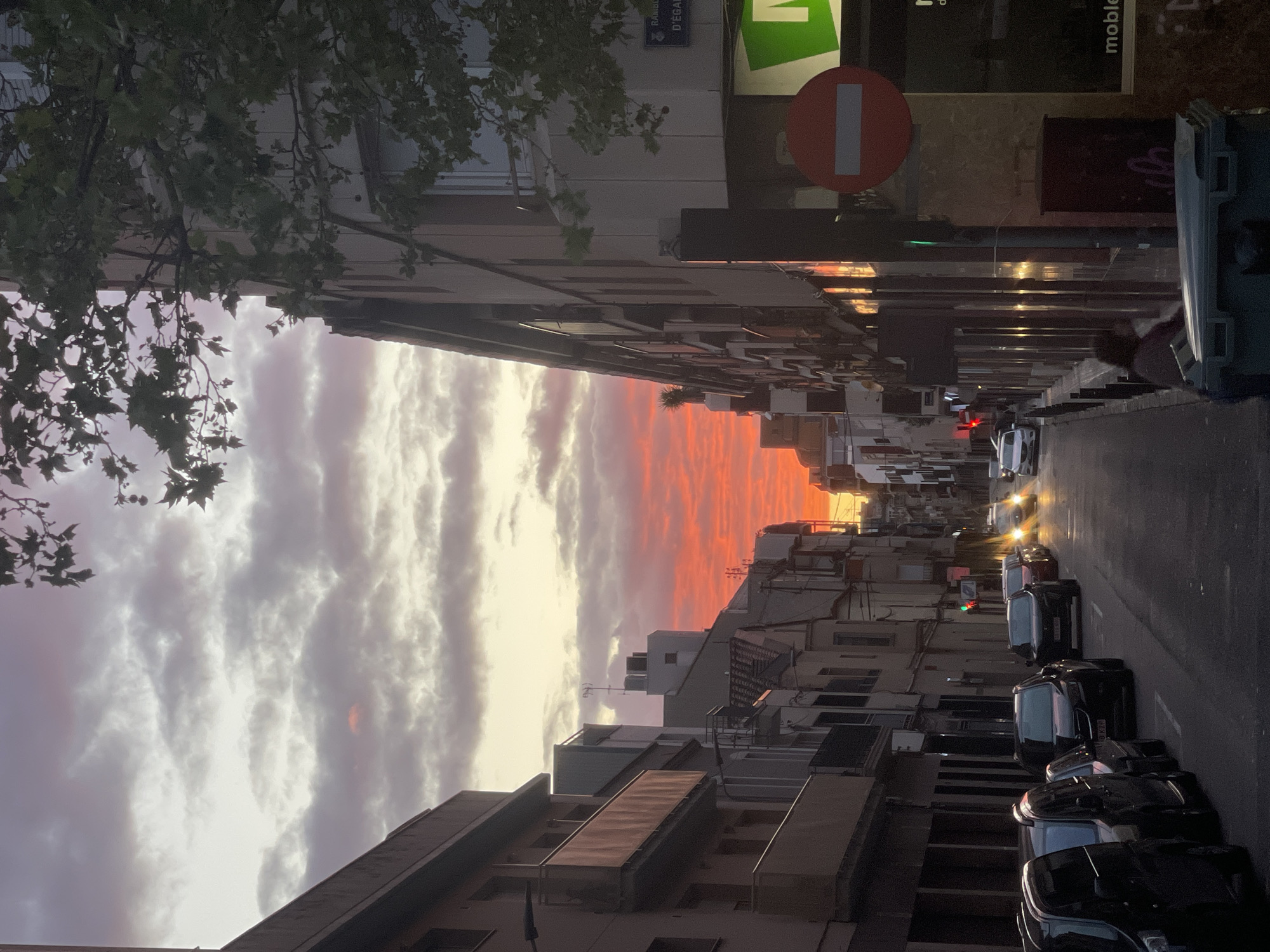 Captured from an elevated vantage point, the photograph presents an eastward-oriented view of a street in Terrassa, Spain, at sunset. The sky displays a gradation of colors, from reddish-orange near the horizon to a mix of white and grey higher up.
The street, lined with several-story buildings, is filled with parked cars on the right side. In the distance, car headlights are visible. Street signs and commercial signage, including a green and white sign and a red prohibition sign, are visible on the buildings. A tree canopy is seen at the top left of the frame.