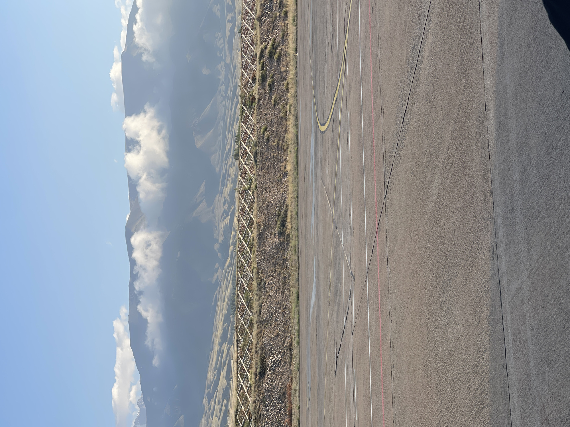This is a daytime, outdoor photograph taken at the Choktal airport in Choktal, Kyrgyzstan. The foreground shows a section of an asphalt tarmac with painted markings, including a yellow curve and parallel white and red lines. Beyond the tarmac, there is a sloped, dry, grassy embankment fortified with a crisscross pattern of wooden or metal structures. Further in the background, a range of mountains is visible, partially obscured by scattered clouds. The sky is blue with some wispy clouds. The scene depicts an airport setting with natural mountain scenery.