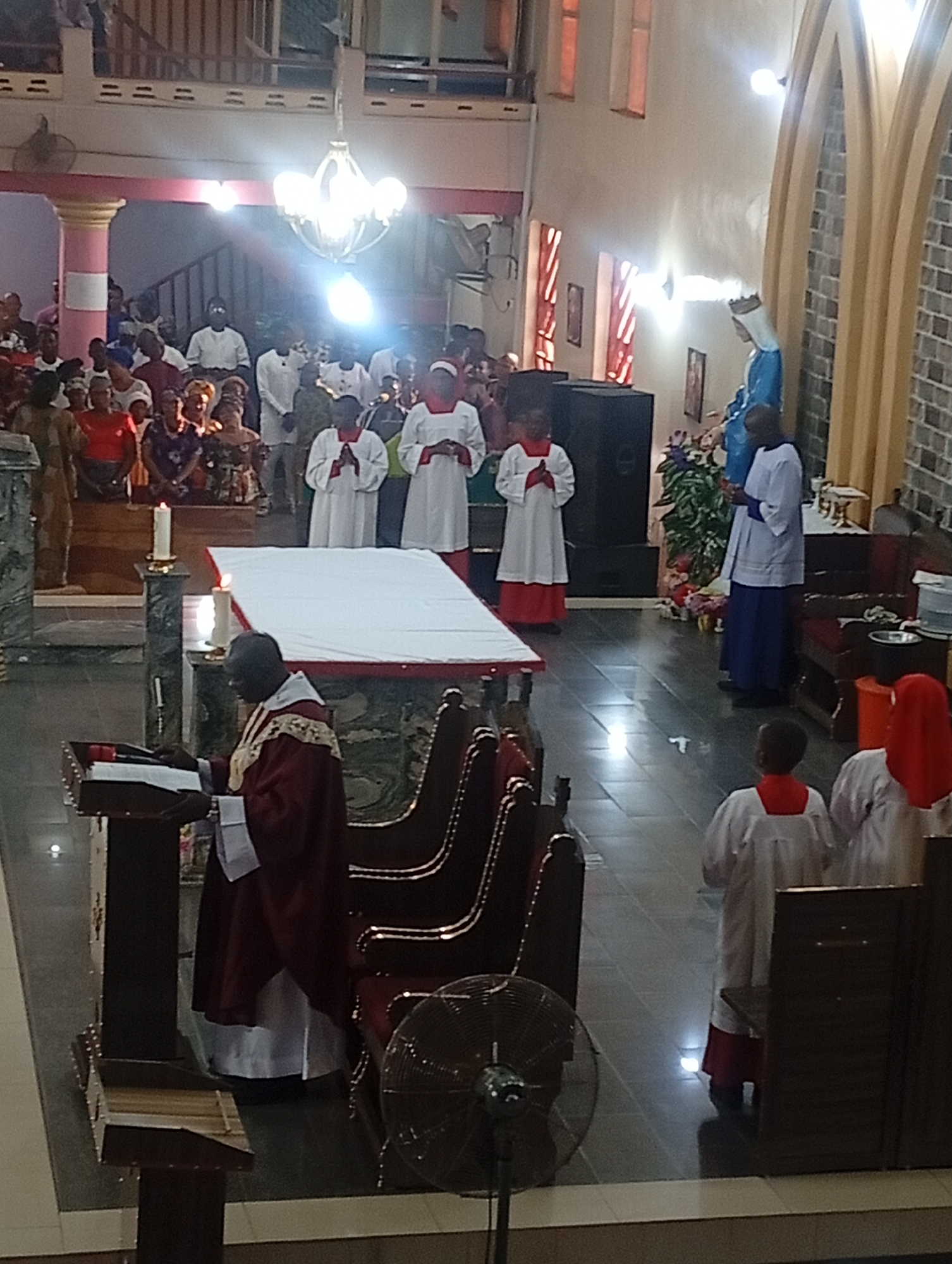 A medium shot shows the inside of a Catholic church. A service is being conducted, with a priest standing at a lectern on the left. A white cloth covers what appears to be a coffin. Altar servers in white robes with red trim stand behind the coffin. Congregants fill the pews in the background. A statue of Mary is in the background near the right side. Arched windows are visible on the right side.
