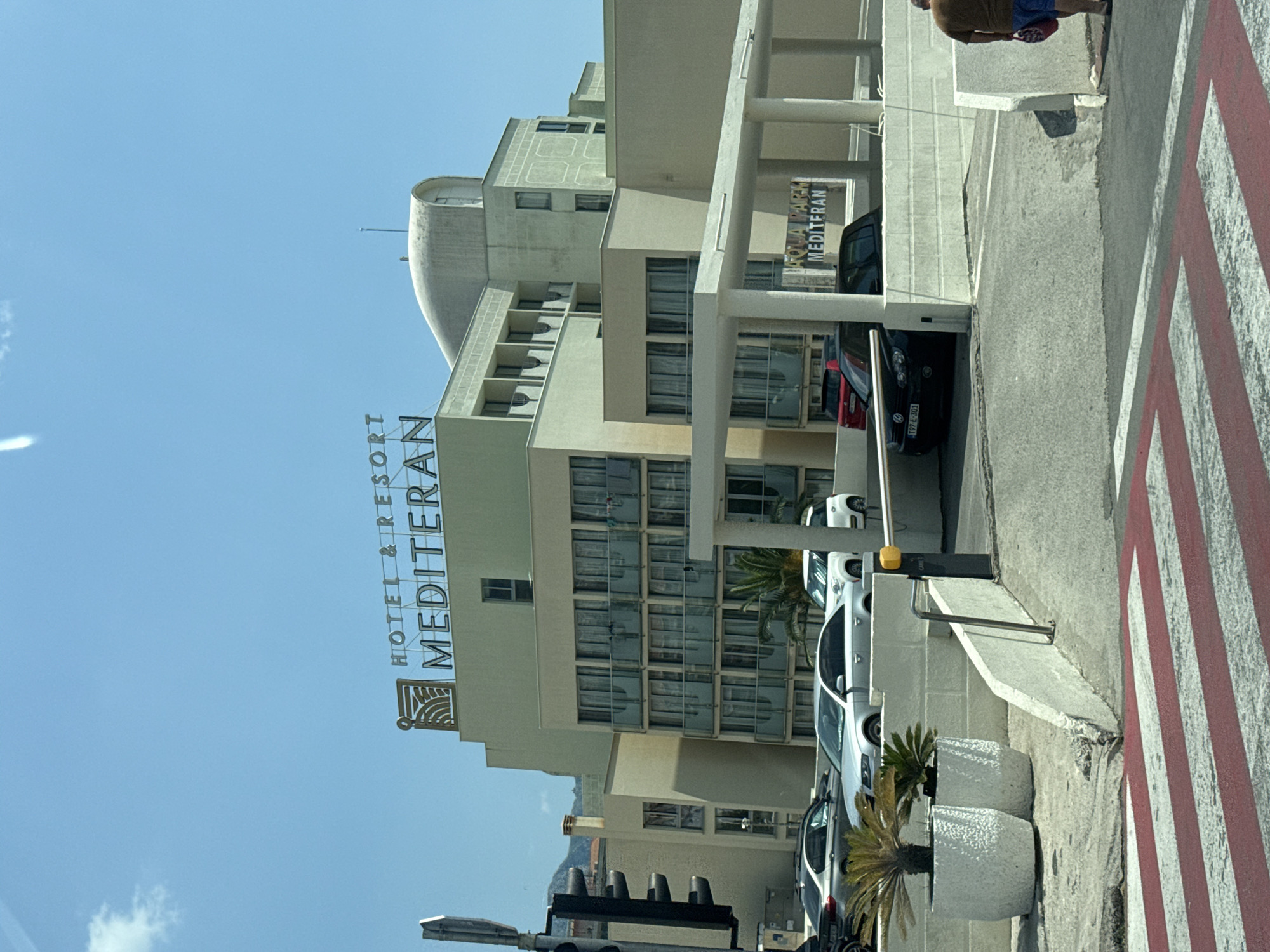 An eye-level, medium shot captures the facade of the Hotel & Resort Mediterran in Bečići, Montenegro, under a clear blue sky. The hotel is a multi-story building with a distinctive curved roofline and a cream-colored exterior. Prominently displayed on the top of the building is the hotel's name in metallic lettering, along with a logo.
In the foreground, a pedestrian crossing with red and white stripes is visible, indicating a street intersection. To the left, a traffic light stands tall. Several cars are parked or driving in front of the hotel, including a silver sedan, a white SUV, and a black Volkswagen.
A white canopy extends from the hotel, supported by pillars, providing shelter over a driveway or parking area. A yellow barrier arm is lowered, suggesting a controlled entry or exit. Two large, white planters with palm-like plants are positioned near the entrance.
A person wearing a brown shirt and blue shorts walks away from the camera towards the right side of the frame, carrying a red bag. The overall scene depicts a sunny day at a coastal resort.