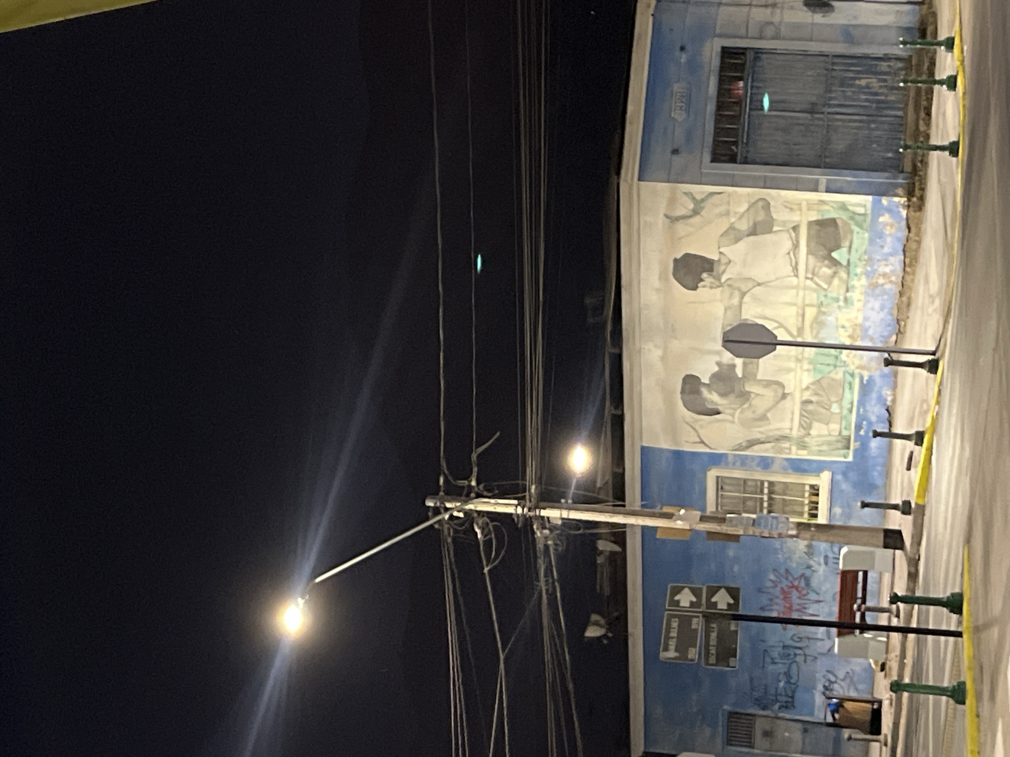 The image shows a street scene at night. A street lamp illuminates a building with a blue facade. The building features a mural depicting figures, possibly people, engaged in some activity. There is also graffiti visible on the lower part of the building. A street sign with arrows and text is present. Power lines run across the sky. The location is in the city of Unknown, Unknown.