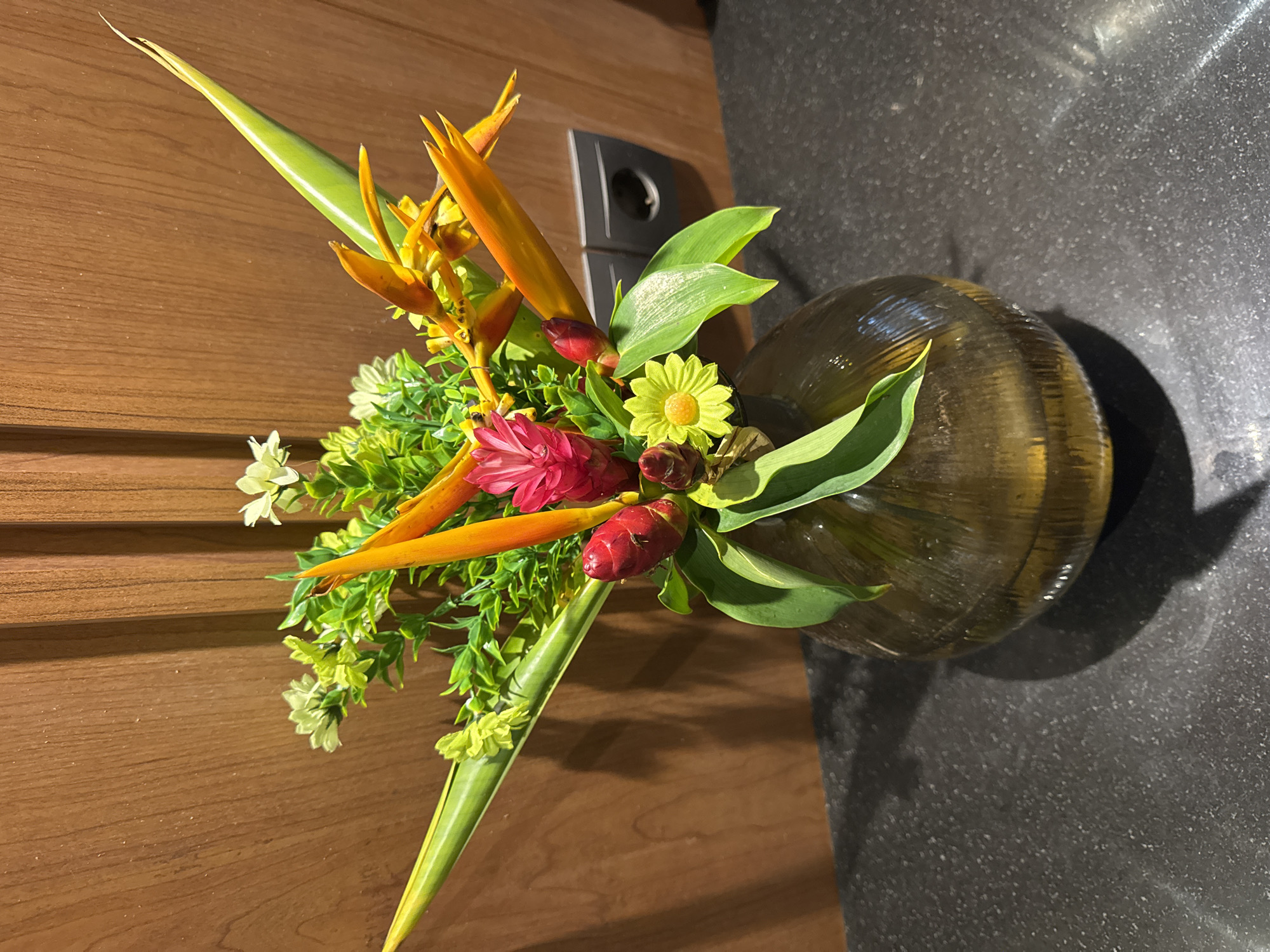 The image, rotated 90 degrees clockwise, displays a vibrant tropical floral arrangement. A dark, glossy vase, with a rounded, bulbous base, rests on a dark, textured surface, likely granite or polished stone. The arrangement features several striking orange and yellow Heliconia blooms, characterized by their elongated, spiky bracts. Interspersed within the bouquet are prominent red ginger flower buds, a small yellow daisy-like flower with a darker center, and delicate clusters of tiny white blossoms. Lush green foliage, including long, pointed leaves and smaller, lighter green sprigs, provides a verdant backdrop to the colorful flowers.
Behind the arrangement, occupying the left side of the image frame, is a warm-toned wooden surface with distinct horizontal paneling, suggesting a door or wall. A dark gray electrical outlet or switch is affixed to this wooden surface. The lighting, likely artificial, illuminates the scene, highlighting the textures and vivid colors of the flowers and the reflective quality of the vase and surrounding surfaces.
The scene depicts an indoor decorative display, exuding an exotic and welcoming ambiance. The specific types of tropical flowers strongly indicate a setting in a warm climate, aligning with the provided location context of Kuta Selatan, Indonesia. There is no human activity or visible text within the frame.