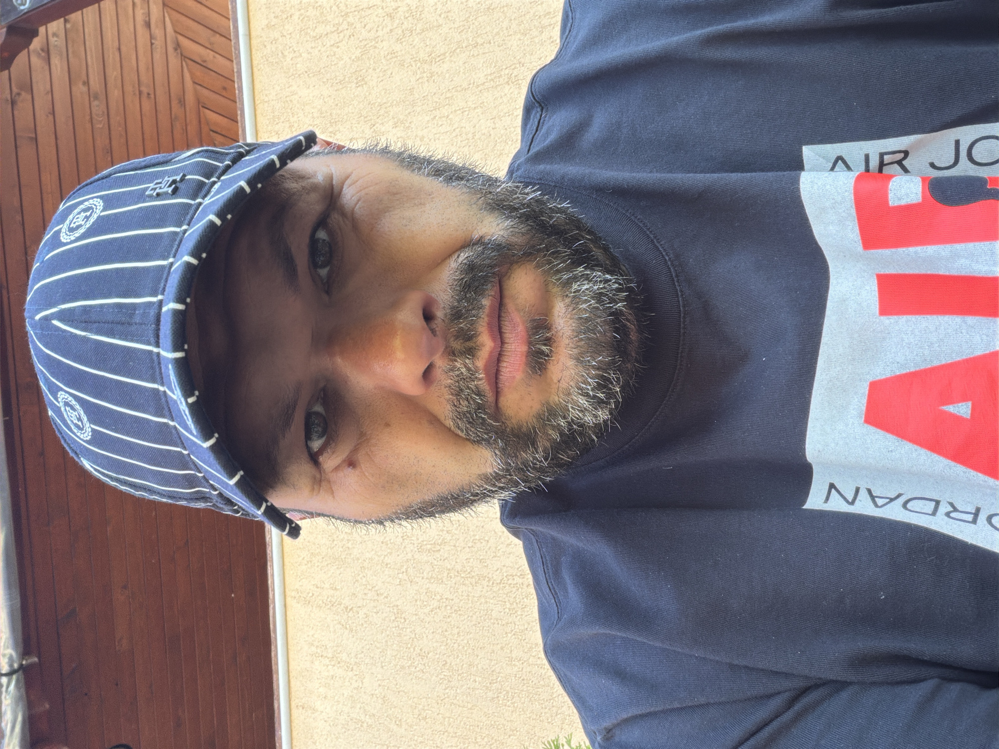 A man wearing a navy blue t-shirt with a "Jordan Air" logo and a navy blue and white striped baseball cap is pictured in a close-up, eye-level selfie. The man has a beard and is looking directly at the camera. The background consists of a tan textured wall and a wooden overhang. The image appears to be taken outdoors during the daytime. There is no specific indication of location within Smolyan, Bulgaria.