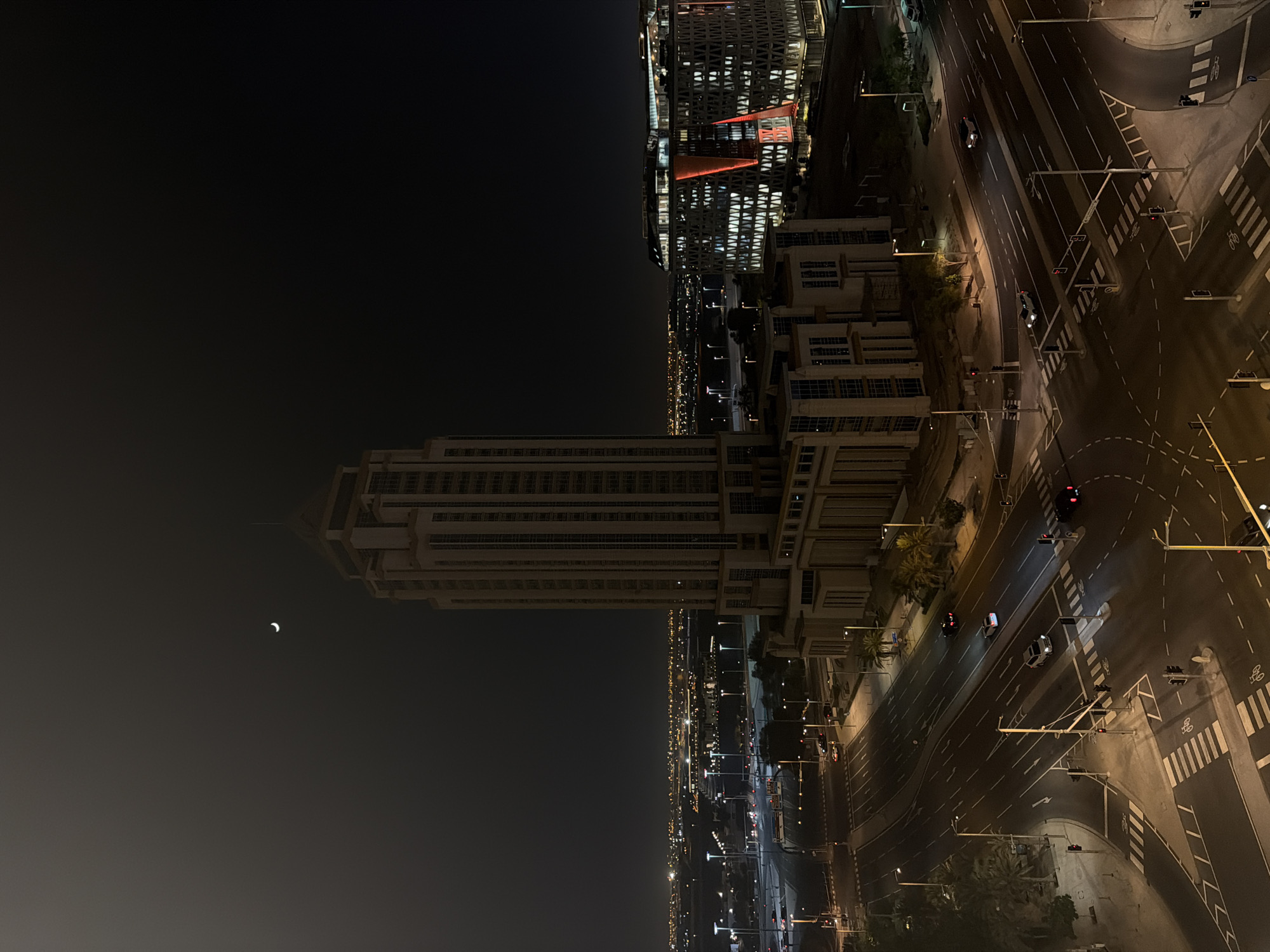 The image is a nighttime aerial view of a cityscape, captured with a vertical orientation. The scene depicts a modern urban environment with tall buildings, roads, and traffic.
In the foreground, a very tall, rectangular building dominates the left side of the frame. Its facade is detailed with horizontal and vertical lines, suggesting a structured architectural design. To the right of this building, and extending further into the frame, are other buildings of varying heights and styles, some appearing more contemporary with geometric patterns and illuminated windows, while others have a more classic, multi-story appearance.
The right side of the image is dominated by a network of roads and intersections. Traffic lights, road markings, and pedestrian crossings are clearly visible. Several cars are present on the roads, some illuminated by streetlights or their own headlights. The roads are illuminated by a combination of streetlights and the lights from the surrounding buildings, creating a vibrant, albeit dark, urban ambiance.
In the dark sky on the left, a crescent moon is visible. The overall impression is one of an active city at night, with ongoing movement and illumination. The time of day is clearly night, given the darkness of the sky and the prevalence of artificial lighting. The weather appears clear, as the moon is visible and there are no indications of rain or fog. There are no discernible people in the image, though vehicles suggest human activity. There is no visible text in the image.