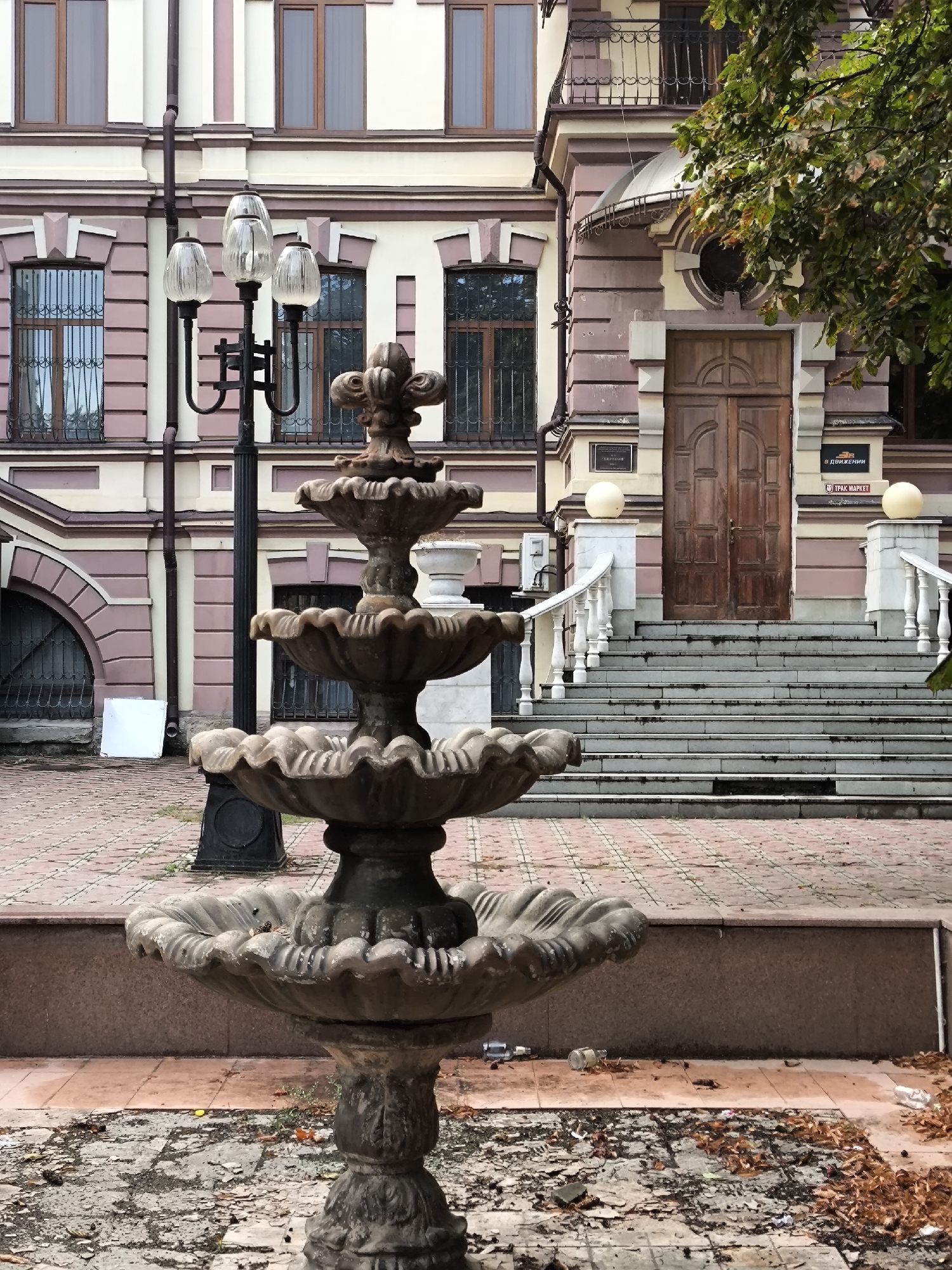 The image depicts an outdoor scene in front of a building with classical architectural elements. In the foreground, a weathered, multi-tiered stone fountain stands prominently. The fountain has a dark, aged appearance and its bowls are ornate. Surrounding the fountain is a paved area with a reddish brick pattern, interspersed with cracked concrete and fallen autumn leaves.
The building in the background features a light-colored facade with pink trim and numerous windows, many of which have decorative iron grilles. A wooden double door serves as the main entrance, accessed by a wide set of stone stairs with white balustrades. A lamppost with three glass shades is positioned to the left of the fountain. A large green tree with some autumn foliage is visible on the right side of the image, partially obscuring the upper part of the building.
The location is identified as being in Yessentuki, Russia. The overall scene conveys a sense of age and possibly disuse, with scattered debris and fallen leaves contributing to a slightly neglected atmosphere.