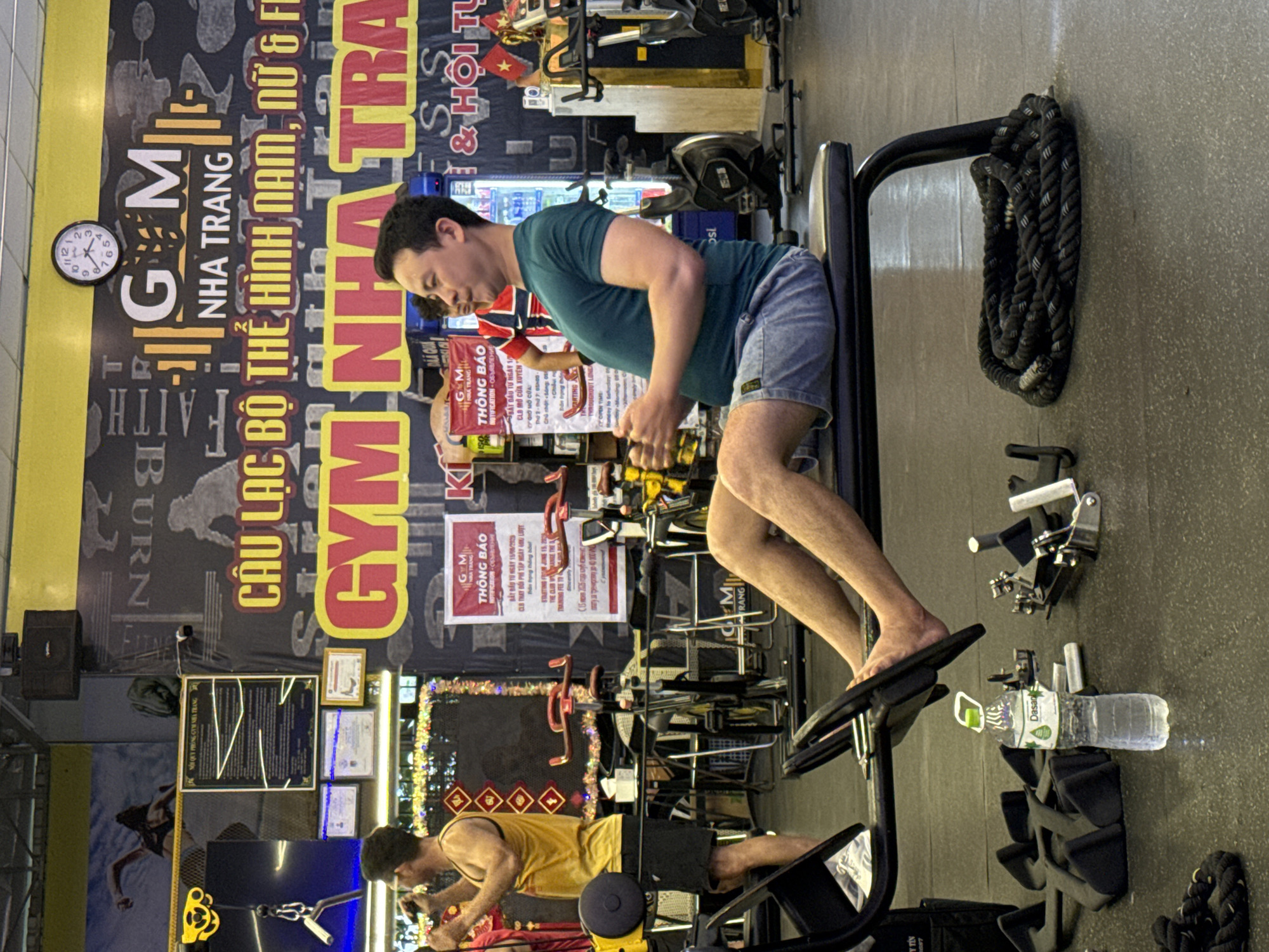 The image shows the interior of a gym in Nha Trang, Vietnam. The gym has a large yellow banner with the text "GYM NHA TRANG" in large red letters. There are several people in the gym, including a man in the foreground sitting on a bench and using a rowing machine. He is wearing a green t-shirt and blue shorts. Another man is standing in the background, wearing a yellow tank top and black shorts. There are also several exercise machines visible, including stationary bikes and a rowing machine. The walls of the gym are decorated with motivational posters and other fitness-related imagery. The overall atmosphere of the gym appears to be busy and active.