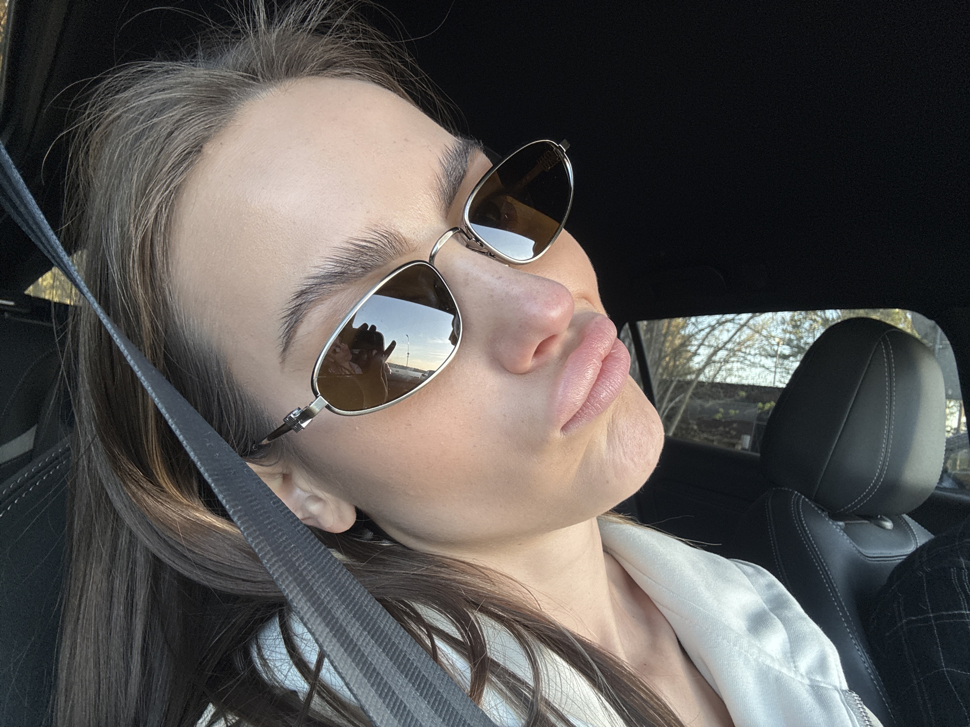 A person with long brown hair is wearing sunglasses and a seatbelt in a car. The person has full lips and is making a pout. The reflection in the sunglasses suggests the car is on a road with trees nearby. The interior of the car is black. It is not possible to determine the location is Białobrzegi, Poland from the image.