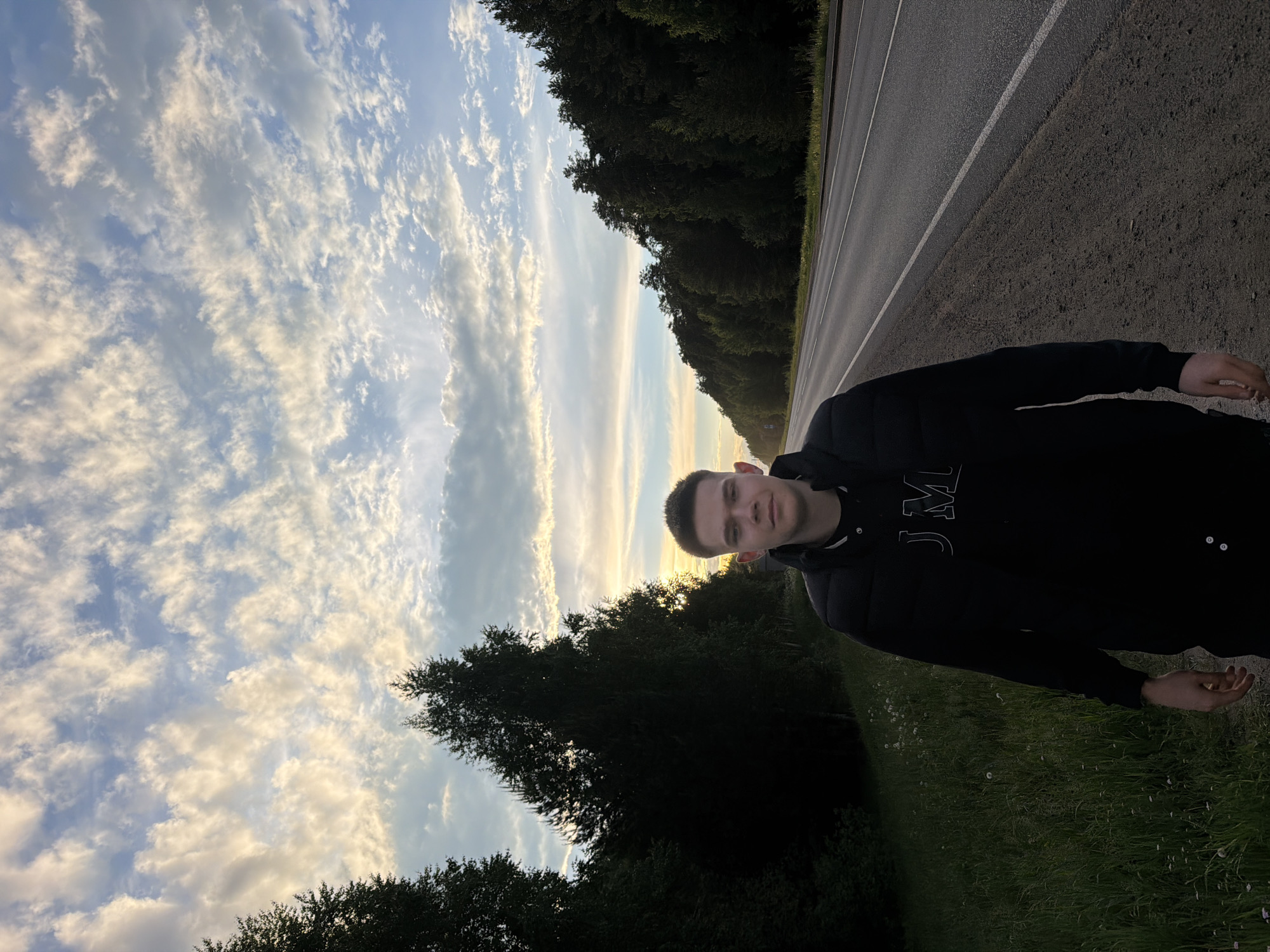 A medium-shot, low-angle photograph shows a young man standing by the side of a road. He wears a dark-colored, puffy jacket. The road runs horizontally across the upper portion of the frame, bordered by a dense line of trees. The sky above is filled with textured clouds, indicating either sunrise or sunset, with the horizon appearing to be the city of Chaikovsky, Russia. The lower portion of the frame consists of green vegetation.