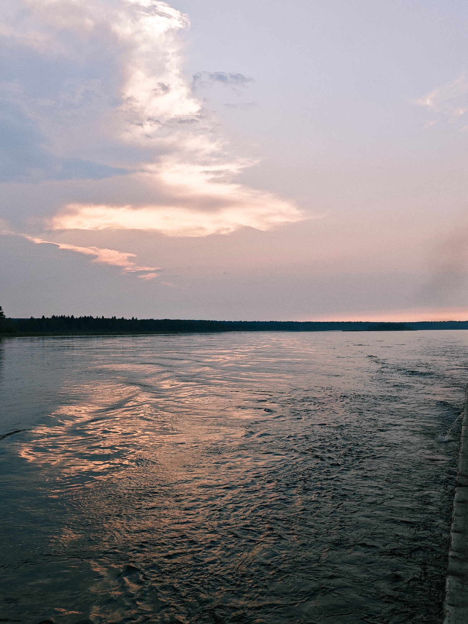 A long shot, taken from the perspective of someone on a boat, looks out over a wide, dark river in Kazachinskoe, Russia. The water, disturbed by the boat's wake, reflects the soft, pastel colors of the sunset sky. The sky itself is filled with scattered clouds, tinged with pink and orange from the setting sun. A dense line of dark green trees forms the horizon on the far bank of the river, their silhouettes stark against the sky. The scene is serene and tranquil, with no people or specific objects visible other than the natural elements of the river, trees, and sky.