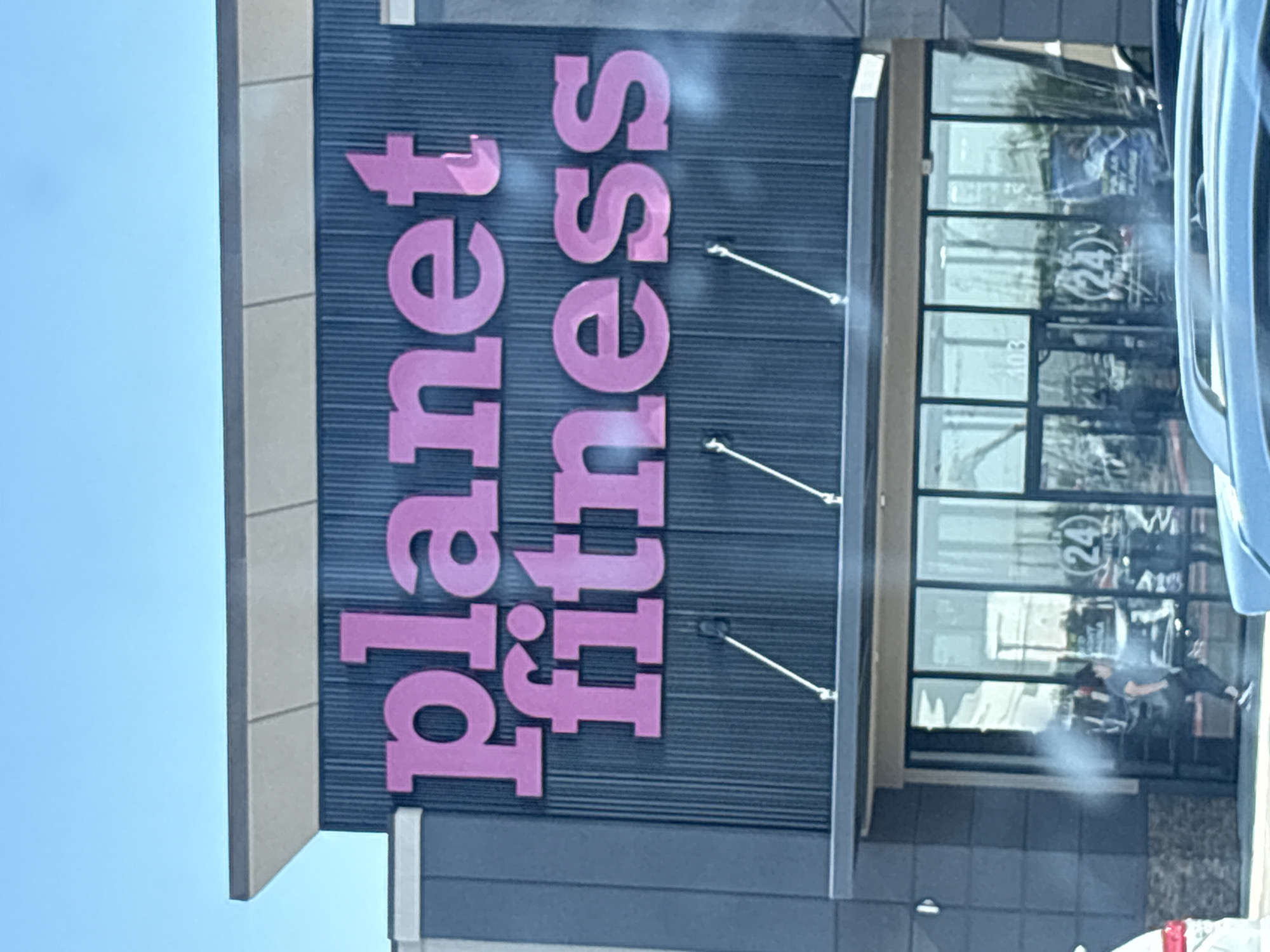 The image shows the exterior of a Planet Fitness gym. The gym's name is displayed in large pink letters on a dark blue, ribbed facade. The building has a modern design with a grey and tan overhang above the entrance. The entrance itself consists of large glass doors, through which some gym equipment and people can be seen. A person wearing dark clothing is visible walking inside. The gym is located in El Paso, United States of America. The sky above is clear and blue.