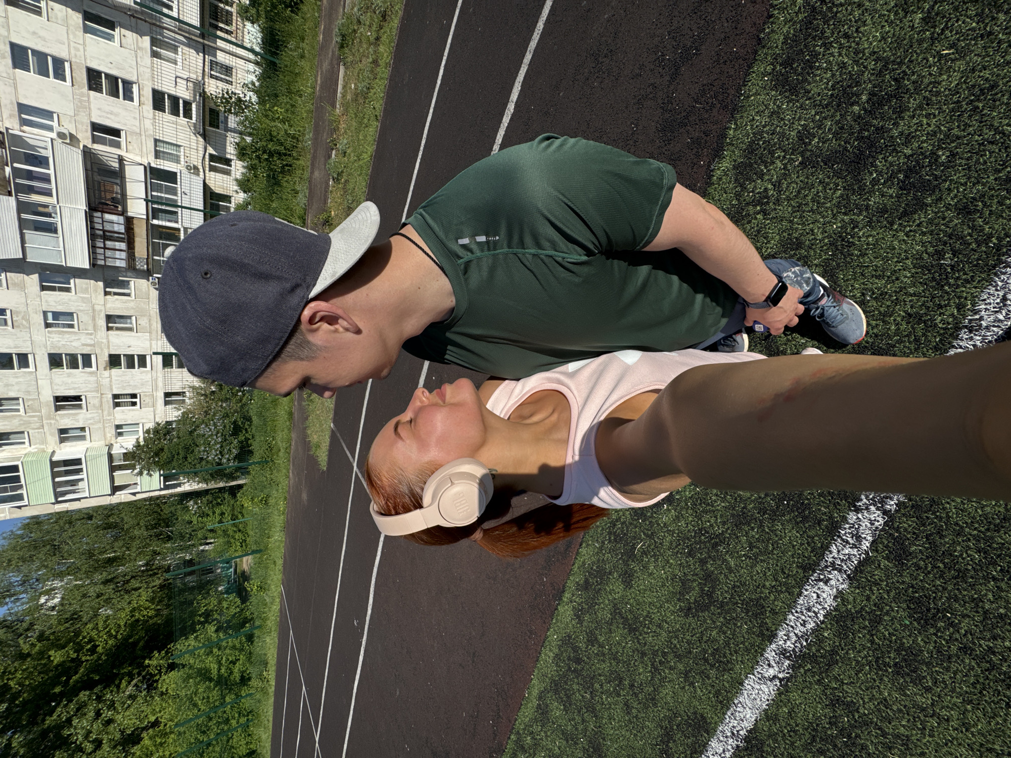 A selfie of two people on an outdoor athletic track in Chaikovsky, Russia is captured on a sunny day. A man wearing a green shirt and dark baseball cap looks towards a woman with red hair and headphones. They stand on a track with white lines on a black surface, next to a green field. In the background is a concrete apartment building with windows.