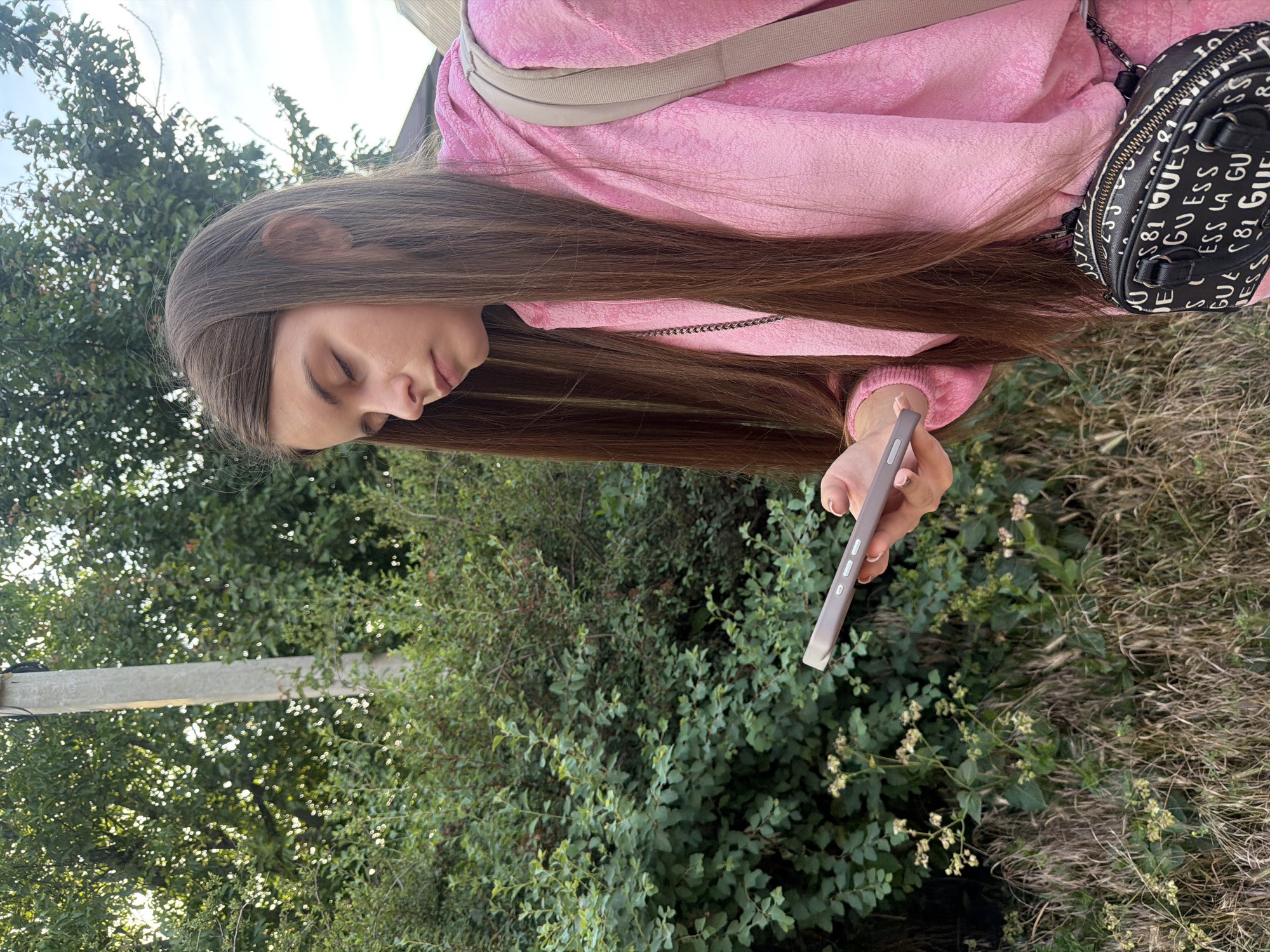 A young woman with long brown hair is looking down at her phone in an outdoor setting. She is wearing a pink hoodie and a black bag with a white logo pattern. The background consists of green bushes and trees, with a utility pole visible to the left. The scene appears to be in a residential area, possibly a yard or a park, in Dnipro, Ukraine.