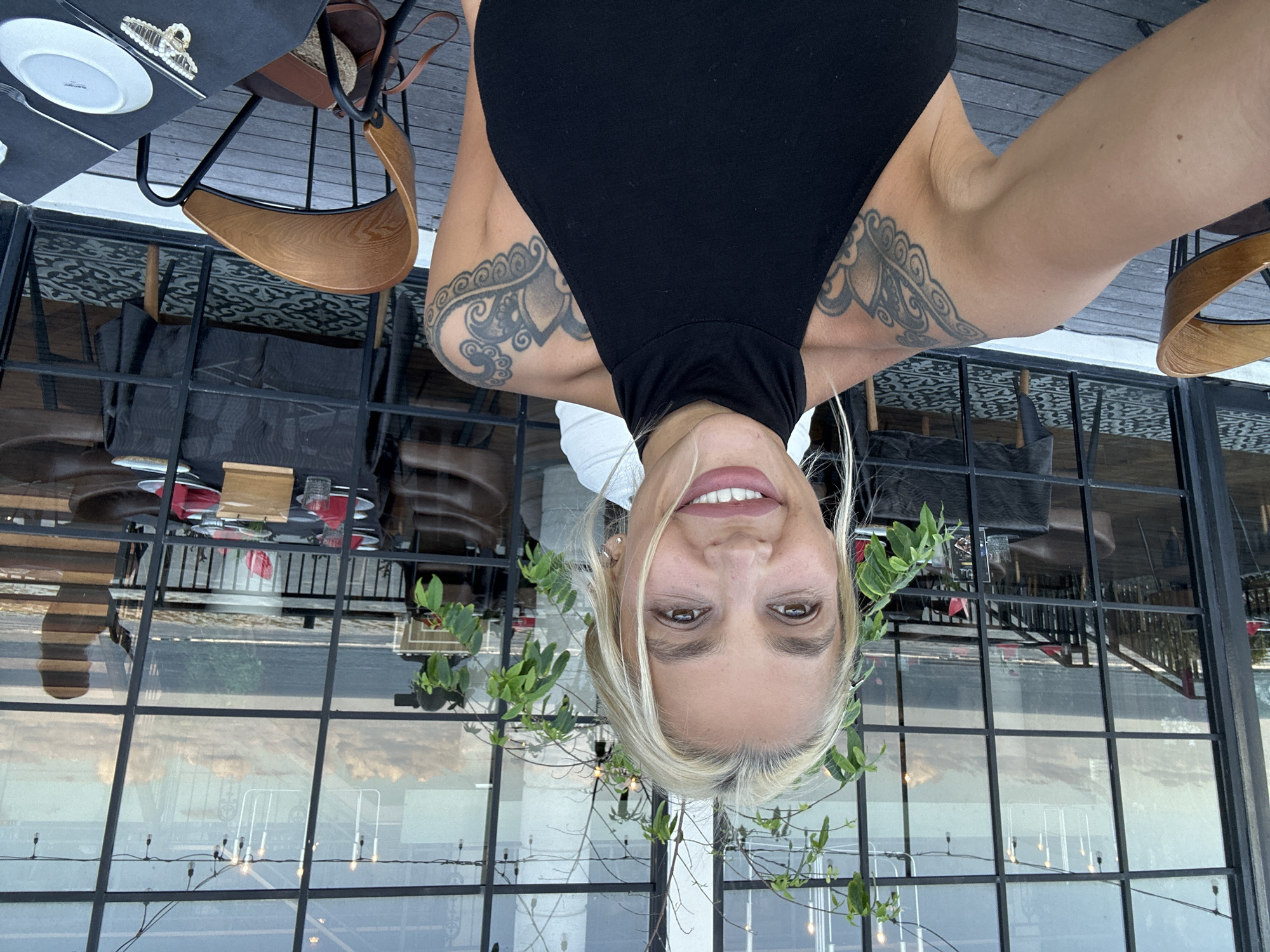 A woman with fair skin and blonde hair, wearing a black sleeveless top, is seen from a close-up perspective. She is facing the camera with a smile. Tattoos are visible on both arms. The image appears to be taken at an upside-down angle.
Behind the woman is a restaurant setting, with tables and chairs visible. The background shows a view of the outside through large paneled windows. Within the window's reflection, a glimpse of landscape with some sky and clouds is visible. String lights are hanging in front of the window. Some plants are placed near the window frame.
Given the description of Kuta Selatan, Indonesia, it can be inferred that the restaurant setting is likely outdoors or has large open windows to capitalize on the tropical scenery. The reflective view visible in the window's reflection shows a hint of the landscape that complements the location's tropical setting.