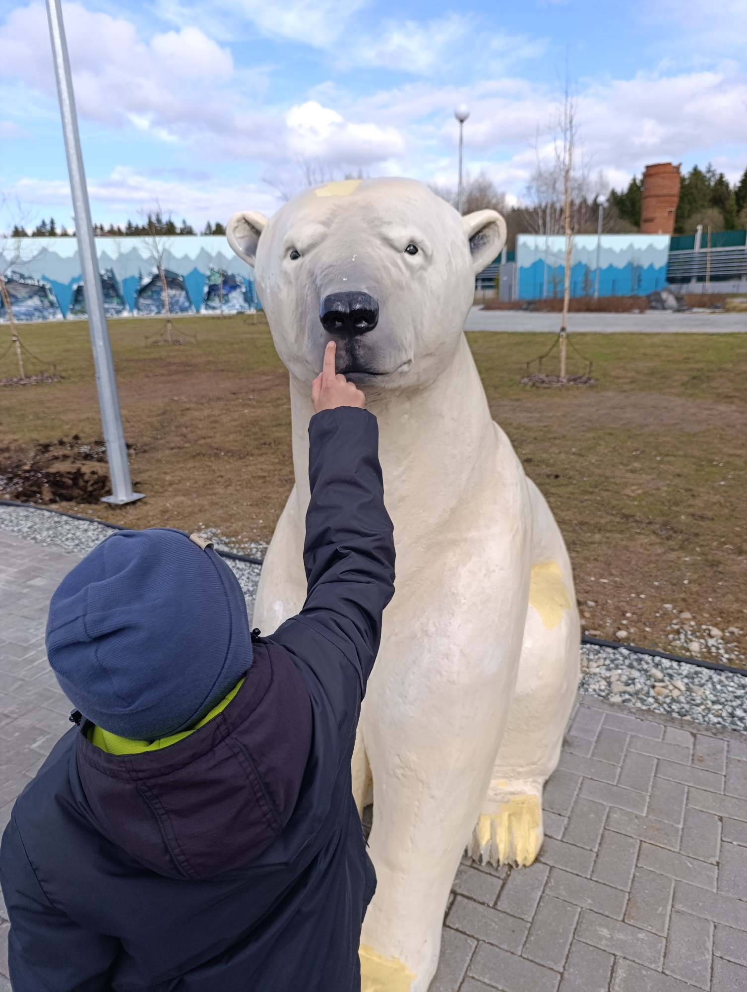 A child in a blue hat and dark blue jacket touches the nose of a large, white polar bear statue. The statue sits on a paved area with brown and gray gravel and short-cut grass in the background. A blue-and-white barrier is visible in the distance. The photo was taken in the city of Perm, Russia.