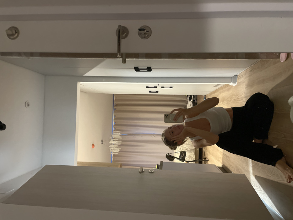A young woman is reflected in a mirror, seated on a wooden floor. She's wearing a white tank top and black pants, holding a smartphone, seemingly taking a selfie. The background shows a bedroom with a beige curtained window, a floor lamp, and a bed. The room has white walls, with a set of white cabinets above her reflection. The mirror's reflection creates an elongated perspective.