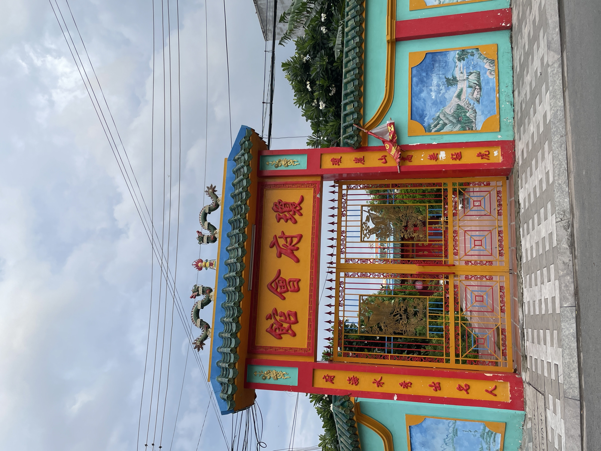 The image captures a vibrant and ornate gate, likely belonging to a Chinese temple or assembly hall, under an overcast sky. The gate is brightly colored, primarily in yellow, red, and teal-blue, featuring traditional Chinese architectural elements.
The central part of the gate is a large yellow panel adorned with prominent red Chinese characters, which read "瓊府會館" (Qióng Fǔ Huì Guǎn), translating to "Hainan Provincial Assembly Hall" or "Hainan Temple/Association." Above this panel, the blue-tiled roof is decorated with several detailed green and red dragon sculptures. The double gates themselves are yellow metal with intricate patterns and feature smaller square cutouts with red and blue designs.
On either side of the main gate, vertical red banners contain more Chinese text. The left banner reads "瓊崖山高拳猛虎" (Qióngyá shān gāo quán měng hǔ), which means "Hainan mountains are high, fists are fierce like tigers." The right banner states "府海水秀出名人" (Fǔ hǎi shuǐ xiù chū míng rén), meaning "The provincial sea water is beautiful, producing famous people." A small red flag with illegible text (possibly "THUỘC") is attached to the upper right section of the gate.
Beyond the gate, green trees and foliage are visible. To the right of the gate, there is a tiled sidewalk with a distinctive gray and white pattern. Overhead, multiple power lines crisscross against the cloudy, bright sky, indicating it is daytime. The overall scene suggests a culturally significant structure in Phan Thiết, Vietnam, likely serving the Hainanese community.