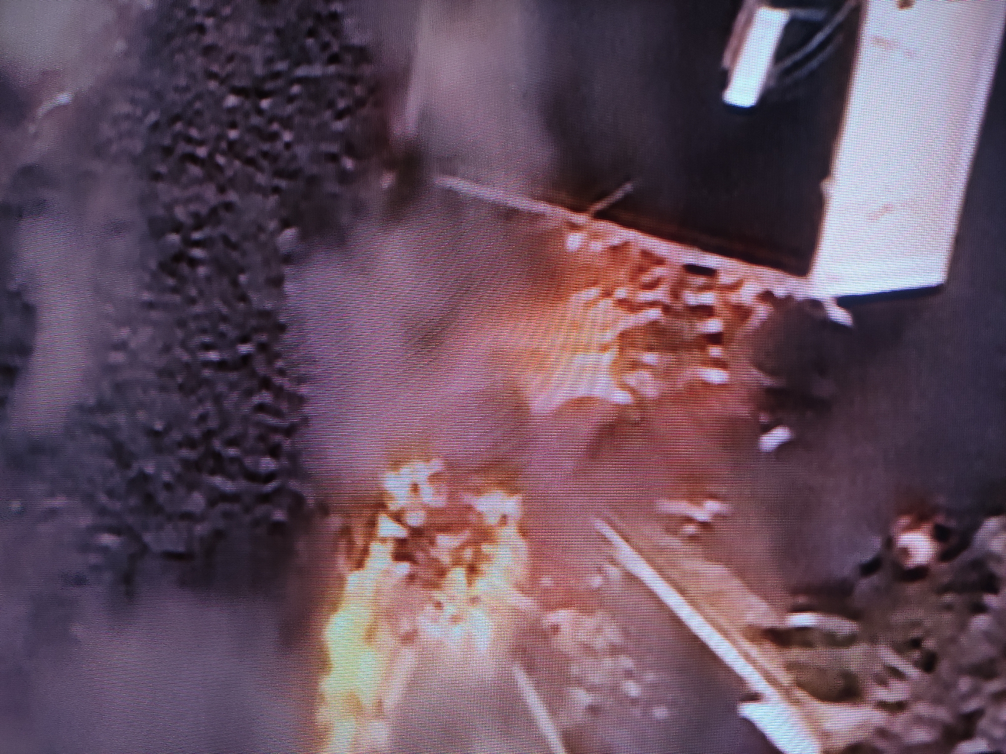 The image, reportedly from Pavlodar, Kazakhstan, depicts a chaotic scene dominated by a large fire and dense smoke. Bright orange and yellow flames are concentrated in the lower central and right areas of the frame, indicating active combustion. A substantial plume of dark grey to purplish smoke rises and spreads, obscuring much of the upper and left portions of the image, suggesting a significant blaze or explosion.
On the far right, a light-colored, rectangular structure, possibly part of a building or an industrial component, is partially visible above the fire. The ground immediately surrounding the flames appears dark and uneven, possibly strewn with debris. No people are visible within the frame, and there is no discernible text.
The primary event captured is a fire, likely a substantial incident given the volume of flames and smoke. The time of day is ambiguous due to the overwhelming light from the fire and obscuration by smoke, but the ambient lighting appears dim. The image quality is low, featuring prominent horizontal scan lines, which suggests it is a photograph of a screen displaying video footage. The scene conveys a sense of destruction and an emergency situation.