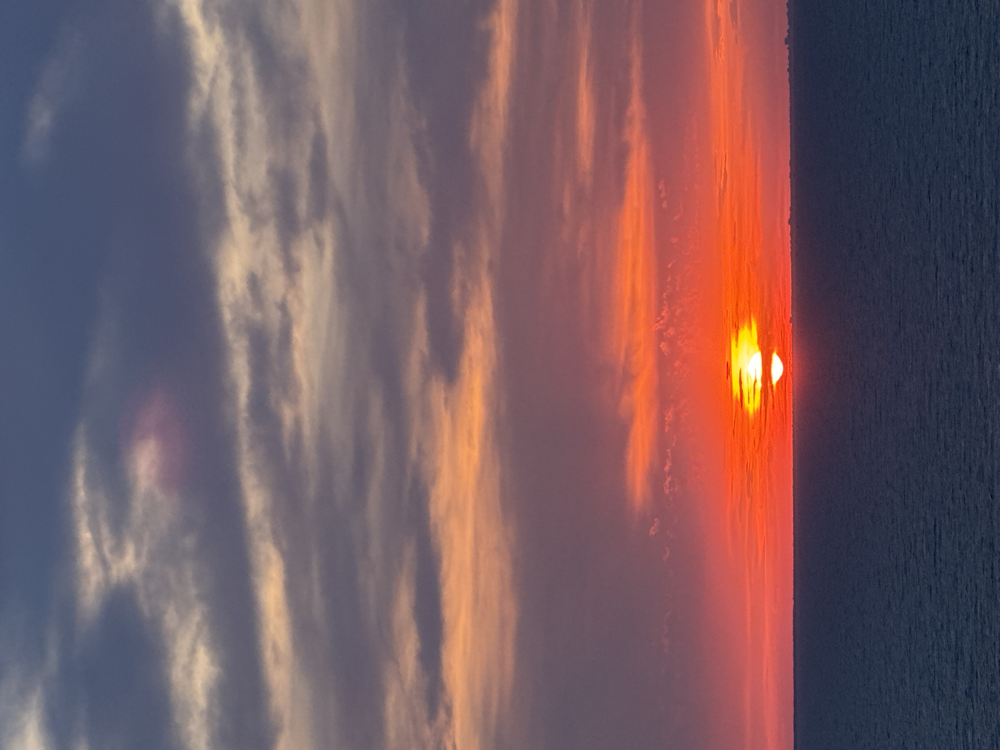 A serene ocean sunset is captured, with the sun partially obscured by clouds, casting vibrant hues of orange and red across the sky. The water below is a deep, calm blue, stretching towards the horizon. A faint outline of a cityscape is visible in the far distance, suggesting a coastal location.