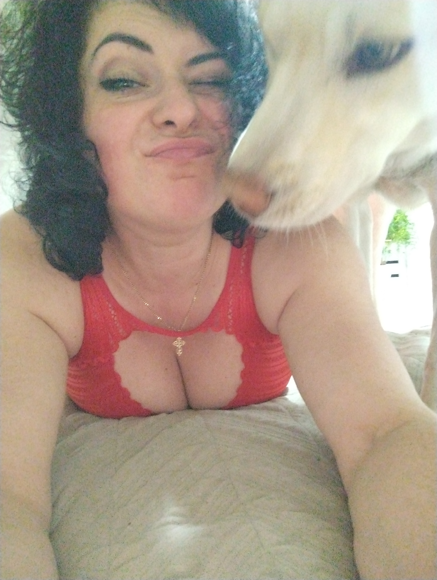 A woman lies on a beige textured surface, possibly a bed, wearing a red top with a keyhole cutout. She is winking at the camera with her right eye closed. She has dark curly hair. A white dog's head is near her face, possibly nudging or kissing her. The environment is unknown. No city is visible.