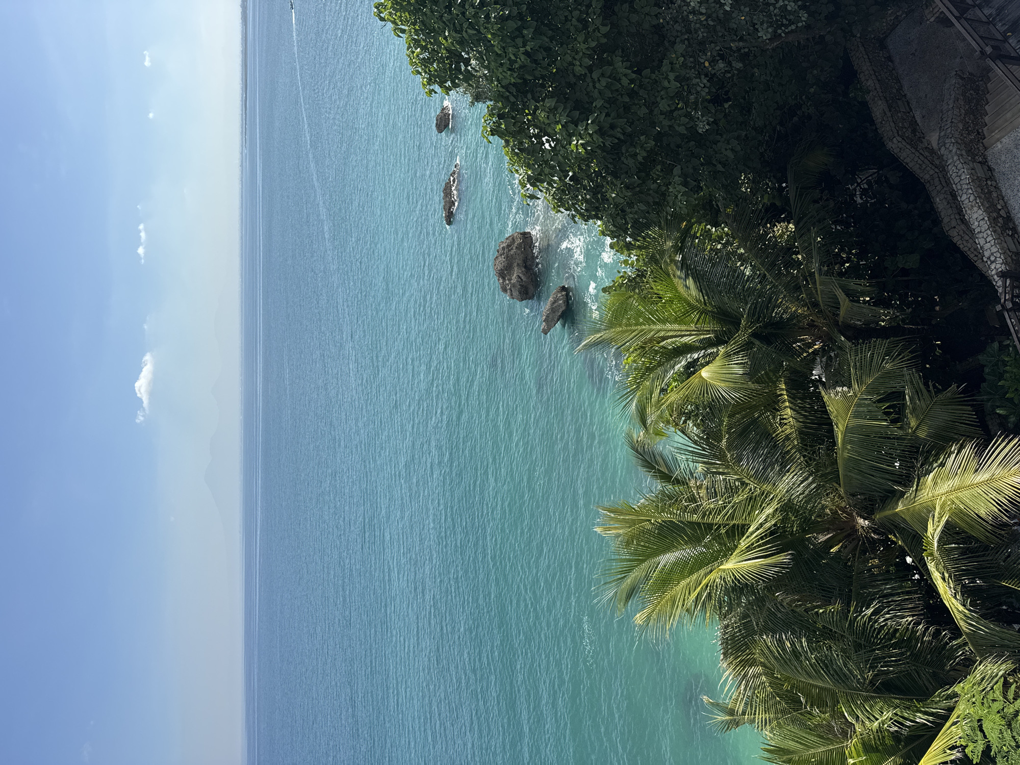 A vertical aerial shot shows a coastal view in Kuta Selatan, Indonesia. Turquoise water occupies most of the frame. Several dark-colored rocks are scattered near the shore, where the waves are breaking. Lush vegetation, including palm trees and other trees, frames the right side of the image. The sky is clear with a single white cloud, and the horizon shows a faint landmass. A boat can be seen in the top left corner. A structure with grey tones is visible in the top right corner.