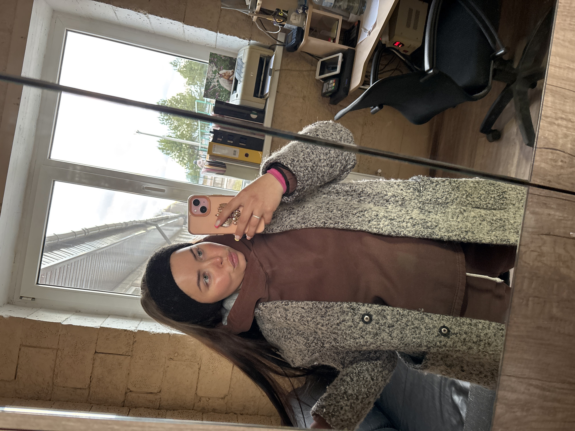 A mirror selfie captures a young woman and the interior of a room, with an outdoor view through a window. The image is oriented vertically, with the top of the room to the left.
The woman, with long dark brown hair and a black headband, is positioned in the foreground. She wears a brown hooded top underneath a mottled grey and green textured coat. Her right hand, adorned with a ring, holds a pink iPhone with a decorative ring grip, used to take the picture. She looks directly at the camera with a neutral expression.
The reflected background reveals a functional workspace with light-colored, possibly brick or stone, walls. To the right, a desk holds a beige printer, a black office phone, a display screen, and various other office items, including yellow and black binders on shelves. A black office chair with a mesh back is also visible. A large window dominates the central and upper background, showing an overcast daytime sky, green trees, and the sloped roof of another building. A thin black line, possibly a reflection or part of the mirror's frame, runs horizontally across the middle of the image. The scene depicts an indoor setting, likely an office, during the daytime in Русский Вожой, Russia.