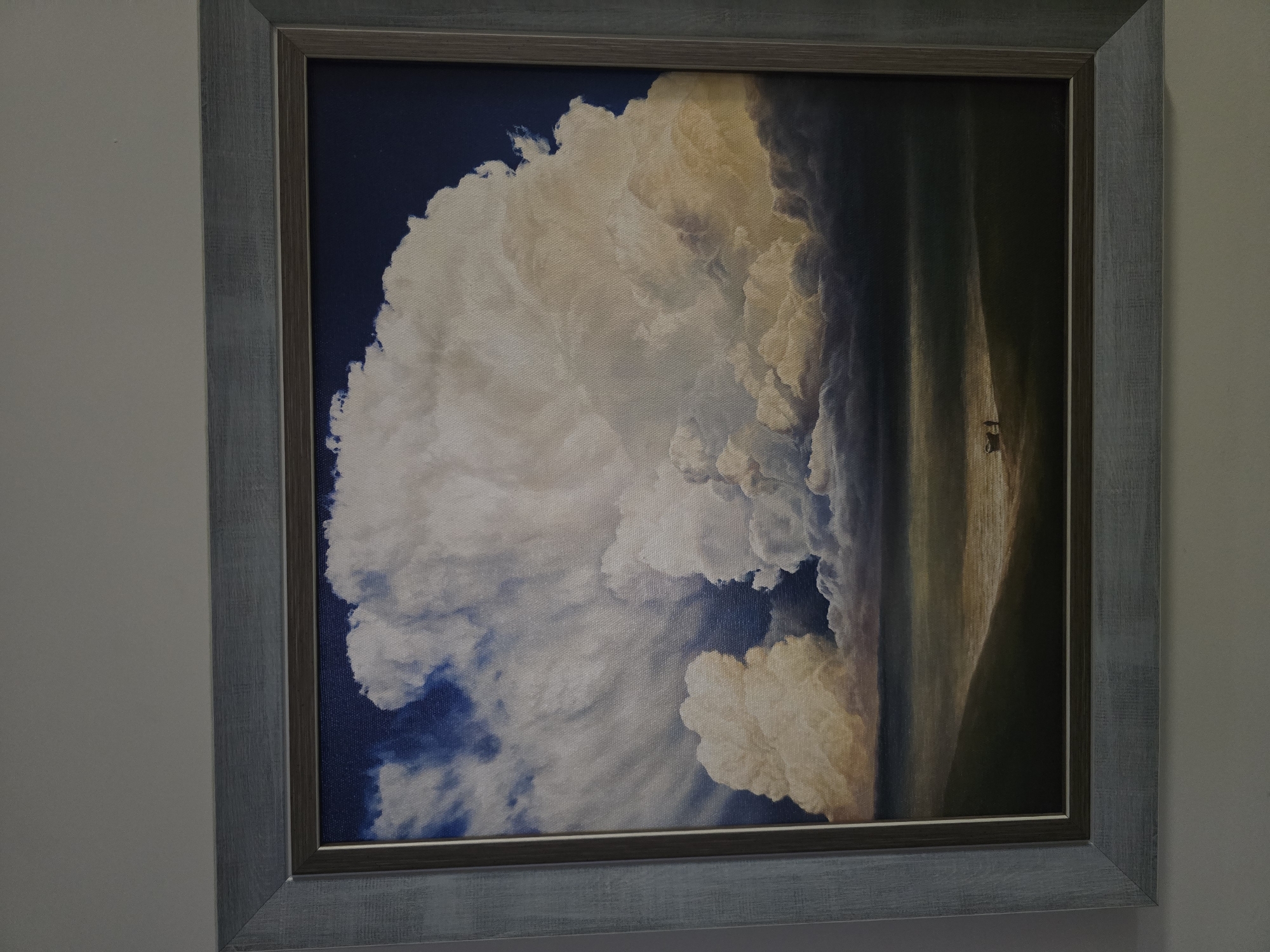 A framed painting is depicted, hanging on a plain, light-colored wall. The artwork portrays a dramatic sky scene dominated by an immense, billowy cloud formation that fills the left side of the canvas. This towering cloud is predominantly white and bright, contrasting sharply with the deep indigo-blue sky visible to its upper left. Its right side is illuminated with warm golden and yellowish tones, suggesting intense, possibly setting or rising, sunlight or a strong internal light source within the cloud.
To the right of the colossal cloud, the painting transitions into darker, more subdued tones, possibly depicting a distant landscape, an approaching storm, or the heavy shadows cast by the cloud itself. Within this dimly lit distant area, a tiny, indistinct dark shape is visible, appearing like a small structure, a vehicle, or possibly a lone figure on horseback, serving to emphasize the overwhelming scale of the cloud. The texture of the canvas is evident across the painting.
The artwork is enclosed in a rustic-looking wooden frame, which is painted in a light greyish-blue color with a subtly distressed or textured finish. No visible text is present in the image.
