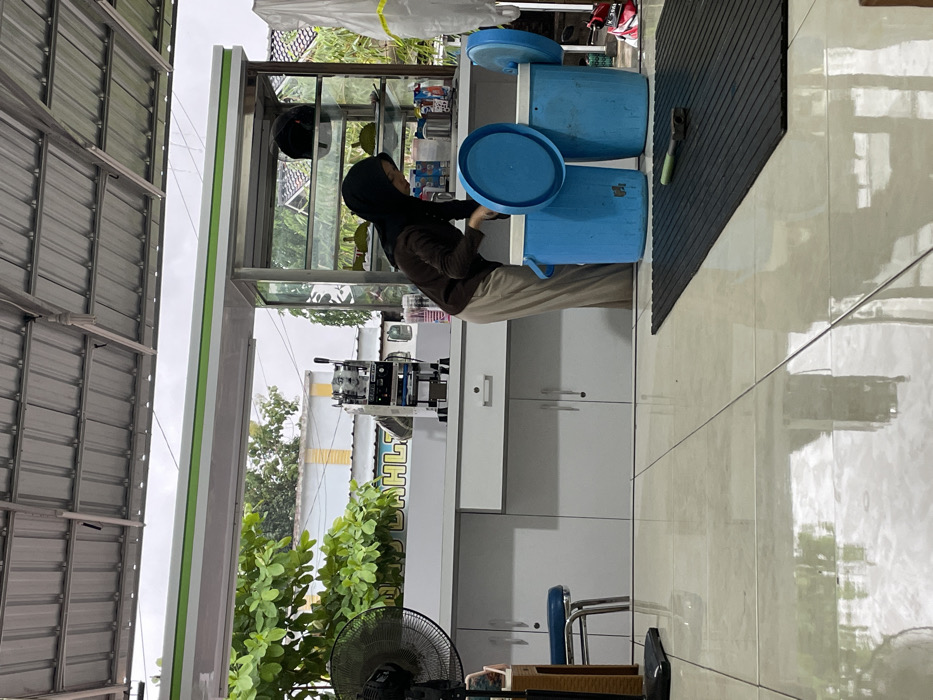 The image depicts a semi-outdoor food or beverage stall in Kayen, Indonesia, under an overcast sky. A single female individual, wearing a dark hijab, brown long-sleeved shirt, and light-colored trousers, is positioned center-right. She is leaning forward, interacting with two large, cylindrical blue insulated containers on the tiled floor. The left container has its blue lid partially open, resting on the rim, while the right container's lid is fully open and leaning against its side.
The foreground features a reflective light-colored tiled floor with a black ribbed rubber mat on the right, on which a small green-handled tool rests. A white counter with multiple cabinet doors and a central drawer extends across the midground. Behind the counter, from left to right, an electric fan is partially visible, followed by a blue stool with chrome legs, a silver cup-sealing machine, and a metal-framed glass display case. The display case contains multiple shelves, holding items resembling pieces of durian fruit and a dark motorcycle helmet on the top shelf. Various plastic cups and bottles are also visible on the counter. Faint text, "DAHLI," is visible on the wall behind the counter.
Above the counter, a white structure with a green horizontal stripe separates the interior from the background. A corrugated metal roof structure with visible beams covers the scene. The background reveals green foliage from a tree on the left, partially obscuring a distant building painted yellow and white. Another building and a white tarp are visible on the far right.