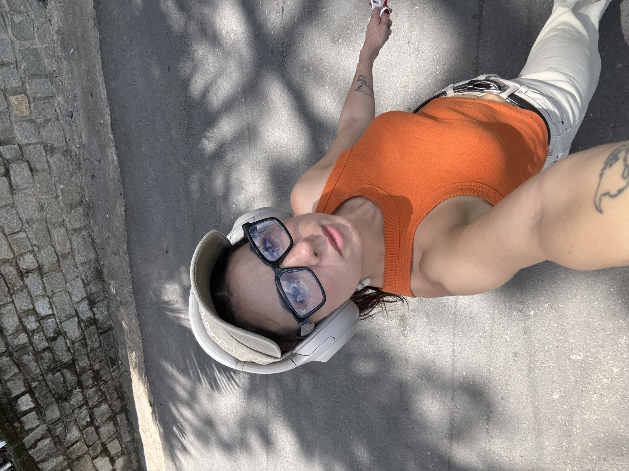 A woman is walking outdoors, holding a leash attached to an unseen pet. She is wearing a baseball cap, large headphones, and glasses. Her outfit consists of an orange tank top and light-colored pants. A stone wall and paved road form the background, with dappled sunlight filtering through palm fronds onto the road. The image is taken from a high angle, looking down at the woman. There is no indication of Da Lat, Vietnam in the visual data.