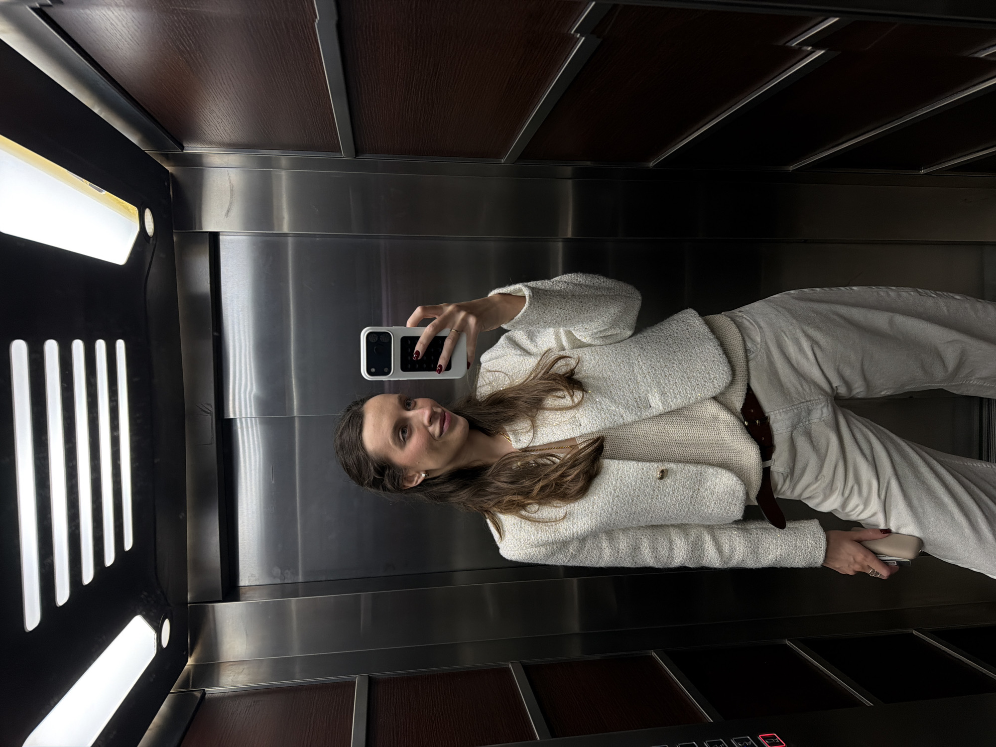 A young woman is taking a selfie in an elevator. She is wearing a white jacket and pants with a brown belt. The elevator has stainless steel walls and brown wood panels. The control panel is visible with red buttons. The location is unspecified, but inferred in the city of Budva, Montenegro, from the user's request.
