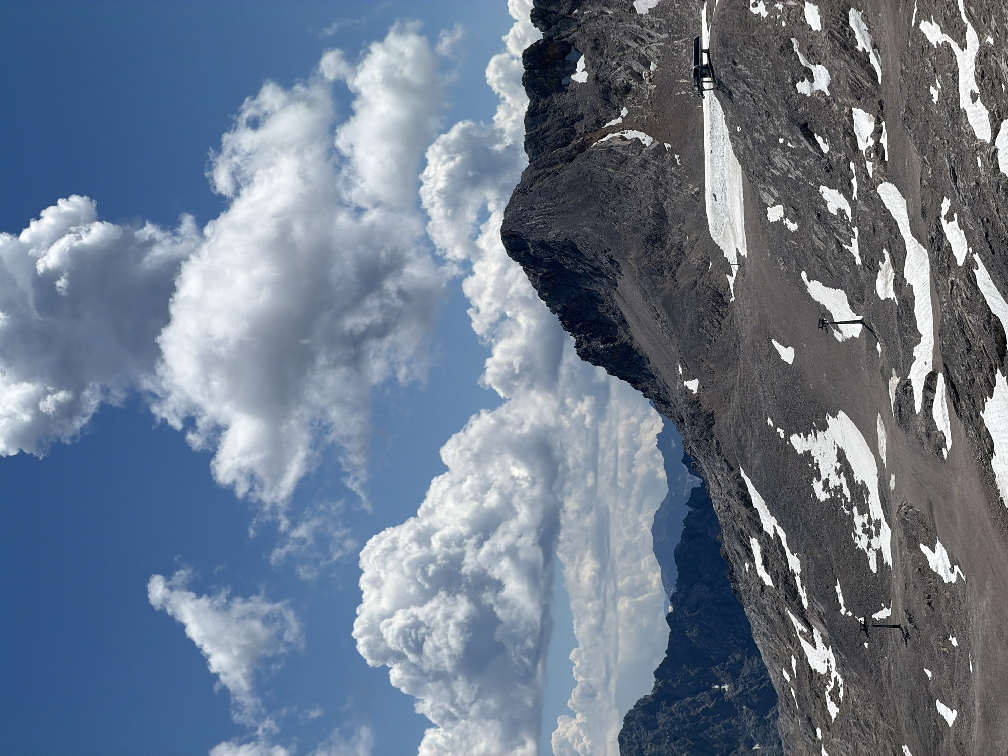 This is a photograph of the Zugspitze, Germany's highest peak, located in Garmisch-Partenkirchen. The image captures a rugged mountain landscape under a bright blue sky dotted with fluffy white clouds. The foreground is dominated by a rocky, barren mountainside with patches of snow scattered across its surface. A ski lift tower is visible on the left side of the frame, and further back on the right, a ski lift station with chairs is perched on the ridge. In the distance, multiple layers of mountains, some appearing dark and jagged, others lighter and softer, recede into the background. The overall impression is one of a high-altitude, alpine environment.