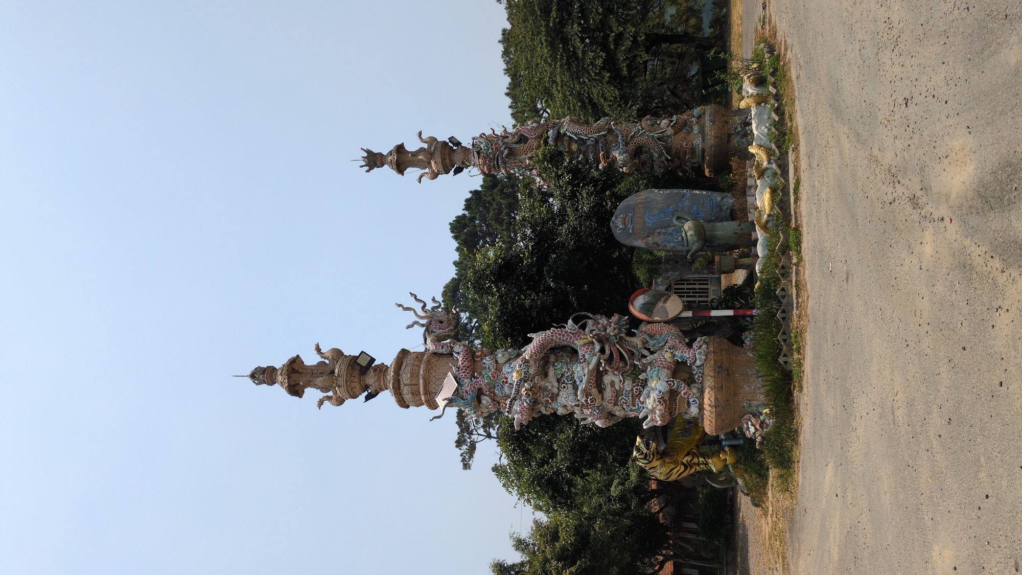 The image shows two tall, intricately decorated columns adorned with dragon sculptures. The columns are located on the side of a road in Lâm Sơn, Vietnam. The columns appear to mark an entrance or boundary to a property or area of interest. Other sculptural elements are visible near the base of the columns, along with a statue. The road is unpaved and runs alongside the columns. The sky is visible above the columns, suggesting an outdoor environment.