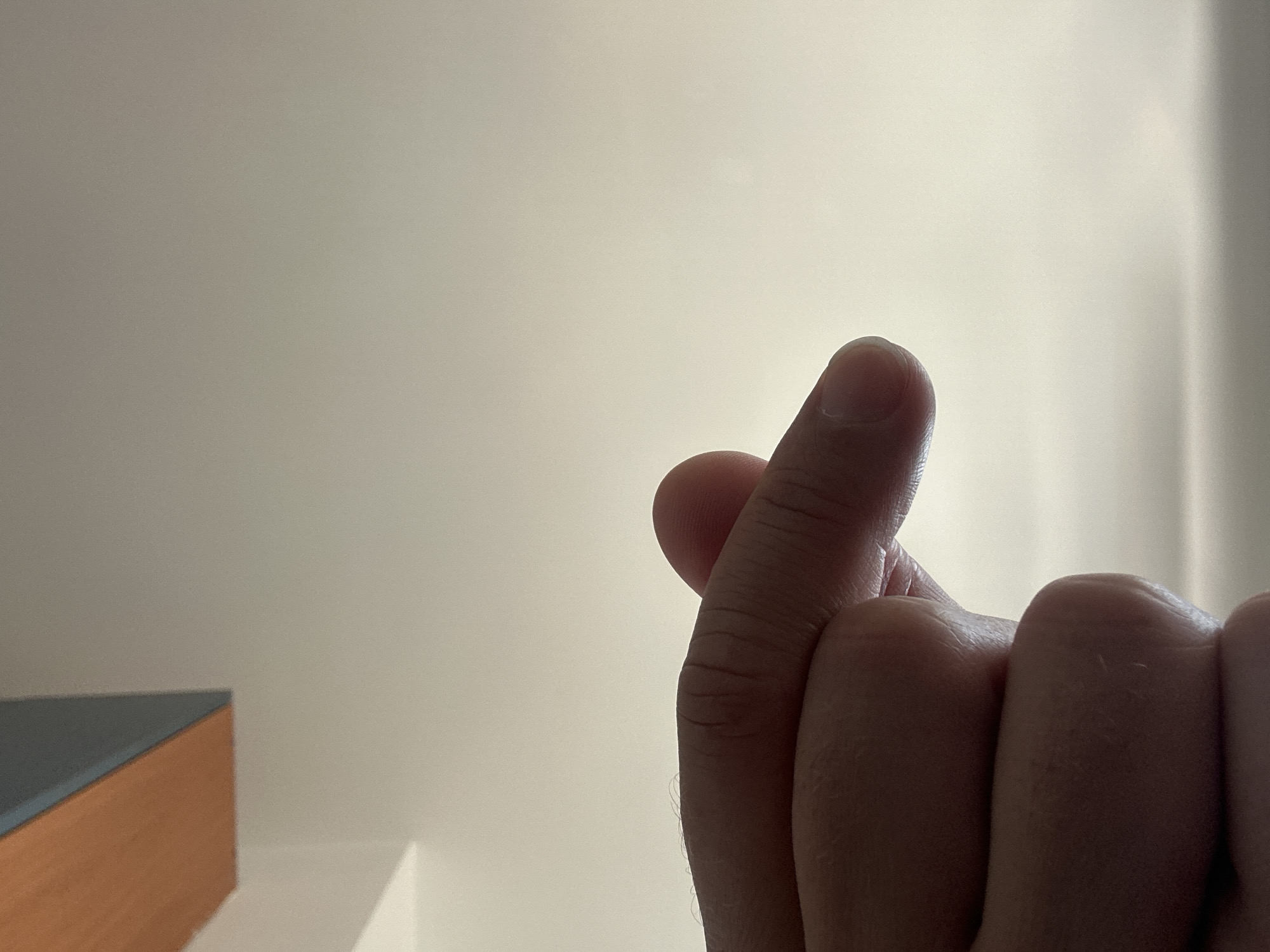 The image features a close-up shot of a person's hand making the "finger heart" gesture. The gesture, formed by touching the thumb and index finger together to create a heart shape, is a popular sign in South Korean culture. The hand is in focus, with the thumb and index finger positioned in the center right of the frame, against a softly lit, neutral background. The lighting suggests it is daytime, possibly indoors, with natural light casting subtle shadows. In the bottom left corner of the image, a portion of a piece of furniture, likely a desk or shelf with a wooden finish and a darker surface, is visible. The overall tone of the image is warm and intimate, due to the gesture and the soft lighting. No text is visible in the image. The context provided places the location in Zvezdara Urban Municipality, Serbia.