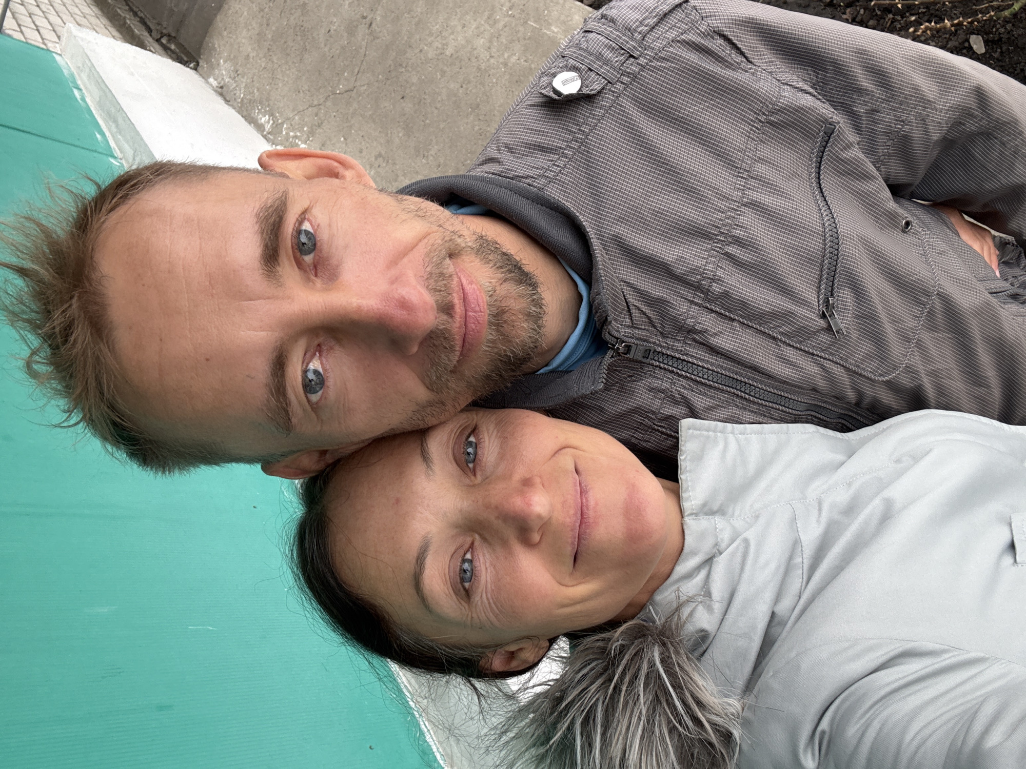 Two people, a man and a woman, are posing for a selfie. The man has short, light brown hair and a stubbly beard. He is wearing a grey zip-up jacket. The woman has dark hair pulled back and is wearing a light grey jacket with a fur-trimmed hood. They are close together, with their heads nearly touching. The background includes a green surface and a concrete area. The location is not discernible as being in Ulyanovsk, Russia, based solely on the visual information provided in the image.