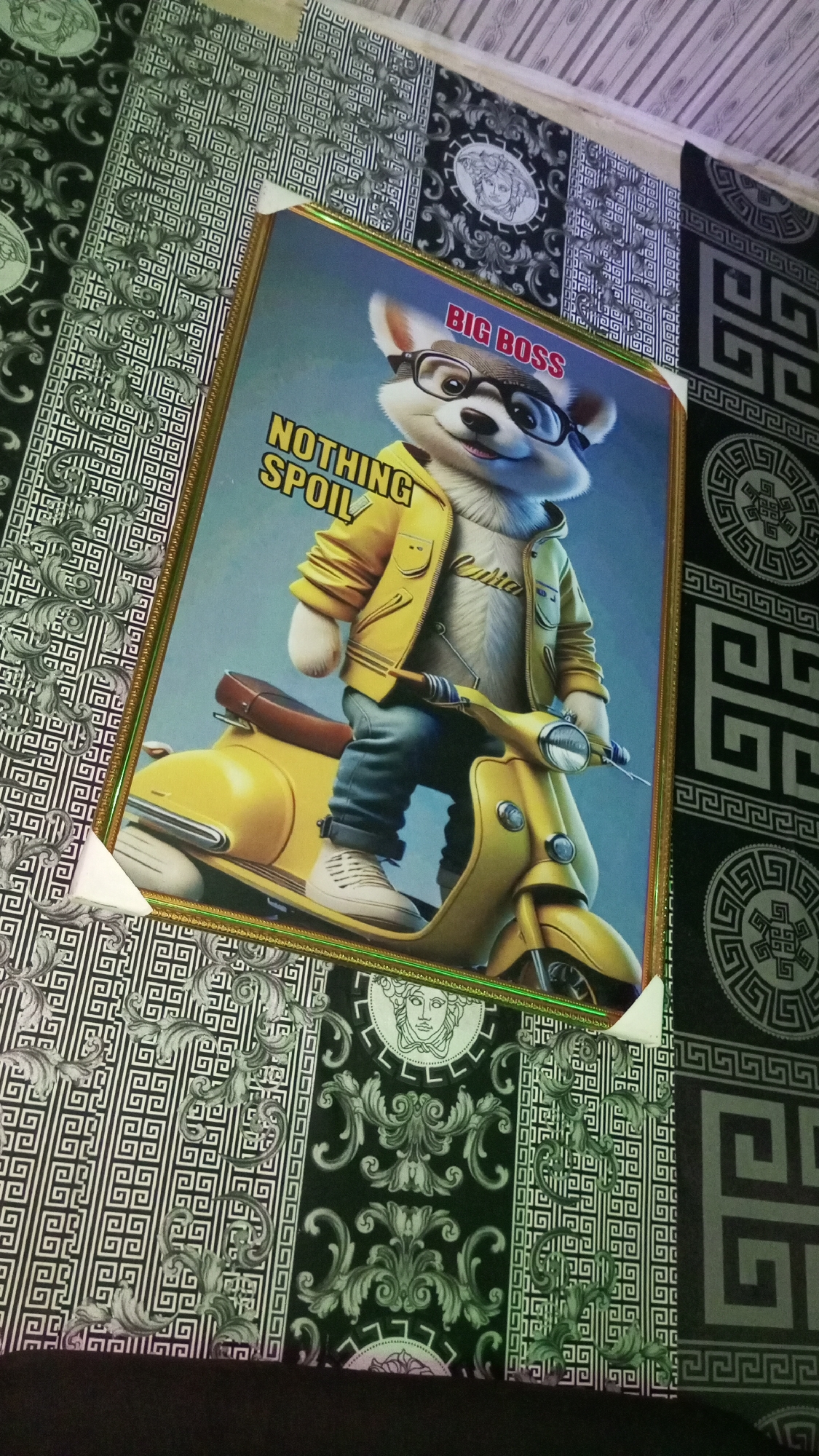 The media file captures a framed picture resting on an intricately patterned surface, likely wallpaper or fabric, in Onitsha, Nigeria. The central subject of the framed picture is a stylized, anthropomorphic white dog-like character, possibly a fennec fox or husky pup, with a cheerful expression. This "big boss" wears black-rimmed glasses, a yellow hooded jacket over a light shirt that reads "Cutie," blue jeans, and white sneakers. The character is depicted posing with a bright yellow scooter against a gradient blue background.
Visible text within the picture includes "BIG BOSS" in red letters at the top right and "NOTHING SPOIL" in yellow letters at the mid-left. The picture is enclosed in an ornate, golden frame, with white triangular corner protectors still attached at the bottom left and top right, indicating it is newly framed or being prepared for display.
The background surface features a detailed and repeating pattern. It alternates between sections of black and white geometric Greek key designs and elaborate black and white baroque-style scrollwork. Circular medallions, each containing a stylized head (resembling the Medusa head often associated with the Versace brand), are interspersed throughout the pattern. The image is taken from a slightly elevated, angled perspective under what appears to be indoor lighting.