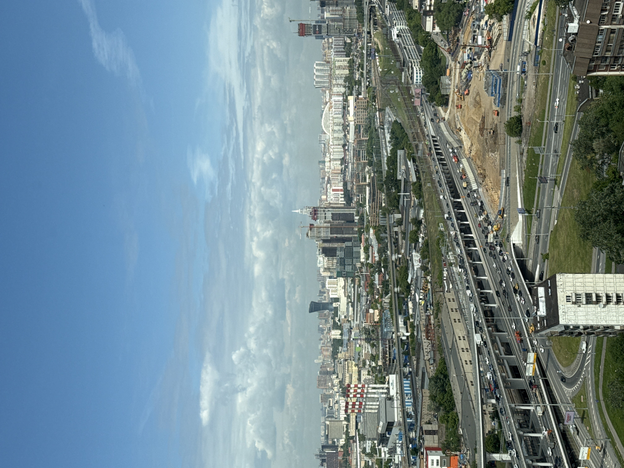 This is an aerial or high-angle view of a sprawling urban landscape, likely in Khimki, Russia, during daytime under a partly cloudy blue sky.
The prominent feature is a complex, multi-level highway system that dissects the city. Numerous cars and trucks are visible on the various lanes and ramps, indicating moderate to heavy traffic flow. Alongside parts of the highway, what appear to be railway tracks are also visible.
The urban environment is dense with a mixture of buildings, ranging from mid-rise to high-rise structures, some appearing residential and others commercial. The cityscape extends far into the background, where more buildings and some cranes signify ongoing development. Notably, one building in the distance features a distinctive red and white checkered facade, while another further back has a dark, modern, and slightly curved design.
Several large construction sites are present, particularly to the right of the main highway, where earthmoving equipment, excavated ground, and unfinished structures can be seen. Cranes are also visible at these sites. Green spaces, including trees and grassy areas, punctuate the concrete and steel of the city.
In the foreground, on a building adjacent to a highway ramp, a sign with the text "FORBET" is visible. The overall scene conveys a busy, developing metropolitan area with significant infrastructure and construction activity.
