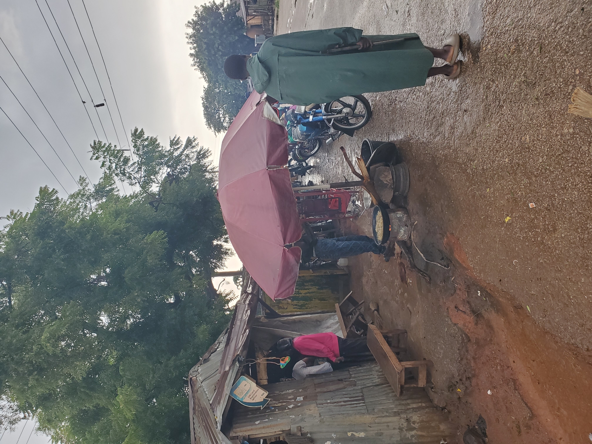 A man in a green rain coat stands facing away from the camera, holding what appears to be a rifle. He is on a wet, gravelly road in what is identified as Barikin Gidar Kanat, Nigeria. To his left, under a large, open, maroon umbrella, are several motorcycles parked. In front of the umbrella, a cooking pot is placed over a fire on a makeshift stove. To the far left of the scene, a small, corrugated metal structure with a thatched roof serves as a shop. Inside this shop, two individuals are visible, one wearing a white t-shirt and the other a pink hooded sweatshirt. A wooden bench is positioned in front of the shop. The environment is overcast, with large trees visible in the background. The ground is muddy and wet, suggesting recent rain. Power lines are strung overhead, with some debris hanging from them.