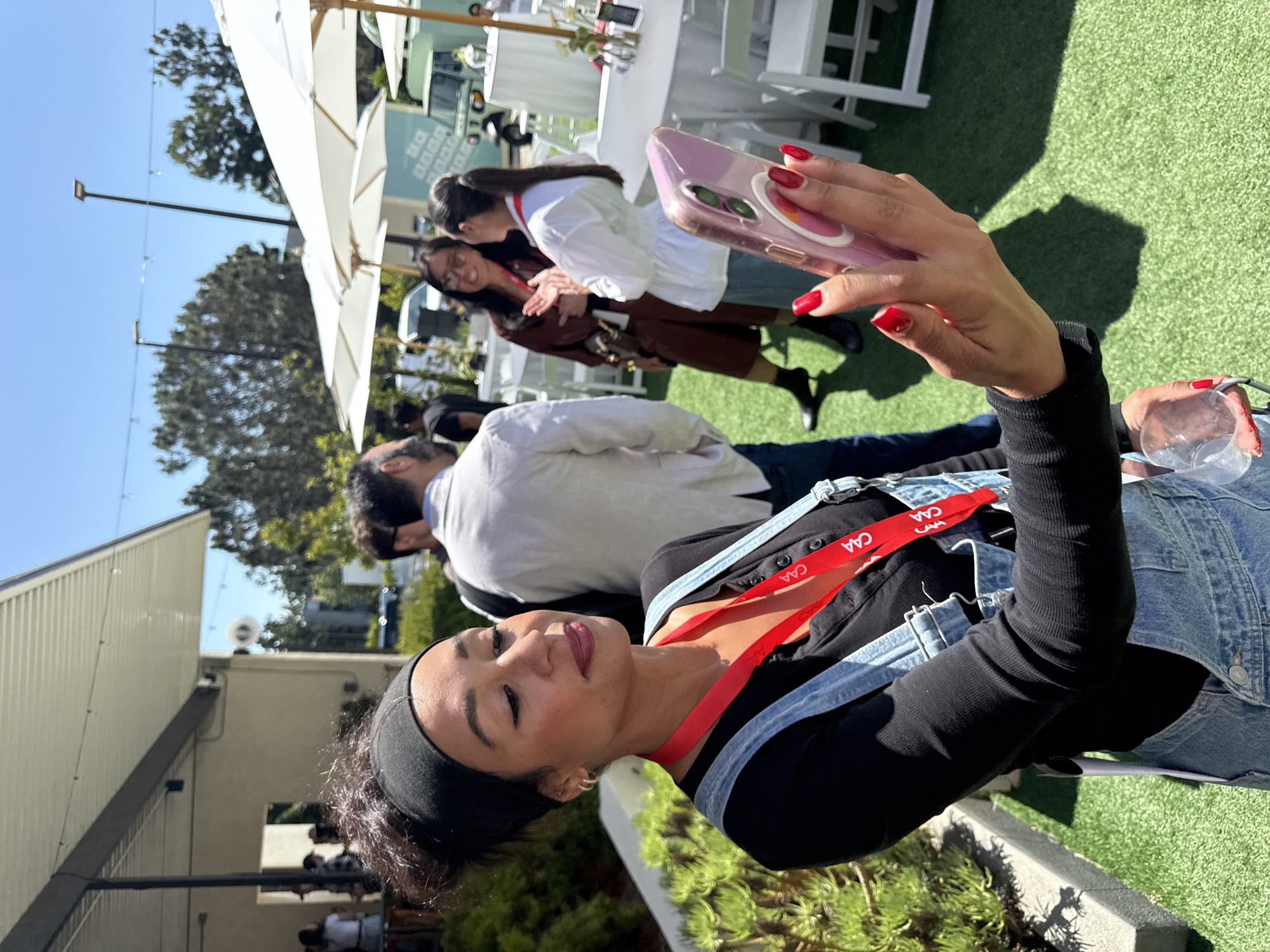 A woman with a black headband and red nails is taking a photo with a pink phone. She is wearing a black long-sleeved shirt and a denim jacket. She has a red lanyard around her neck with "CAA" printed on it. The background shows a sunny outdoor event with white tables and chairs under large white umbrellas. Other people are visible in the background, including a man in a white shirt and two women standing together. The event appears to be taking place on artificial grass. Based on the "CAA" lanyard and the overall vibe, this event could be related to the Creative Artists Agency (CAA). It is impossible to determine the exact location in Los Angeles without further information.