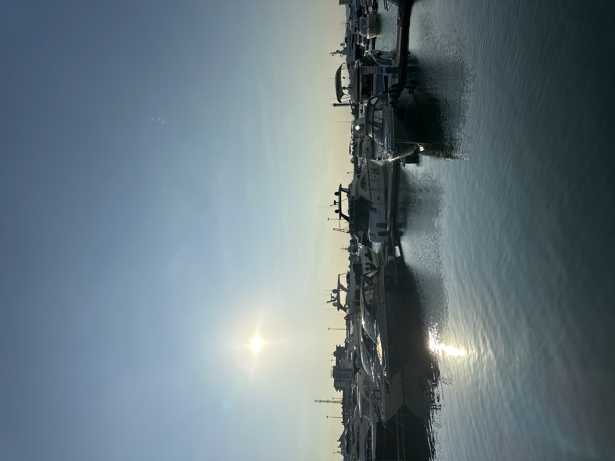 The image shows a marina in Sochi, Russia, filled with numerous yachts and boats docked along piers. The sun is low in the sky, casting a bright glare on the water and illuminating the scene with a warm, golden light. The sky is a clear blue, with a faint haziness towards the horizon. In the background, buildings and port infrastructure are visible, suggesting an active maritime area. The water is calm, with gentle ripples reflecting the sky and the boats. The overall impression is one of a peaceful and scenic harbor, likely during the morning or late afternoon.