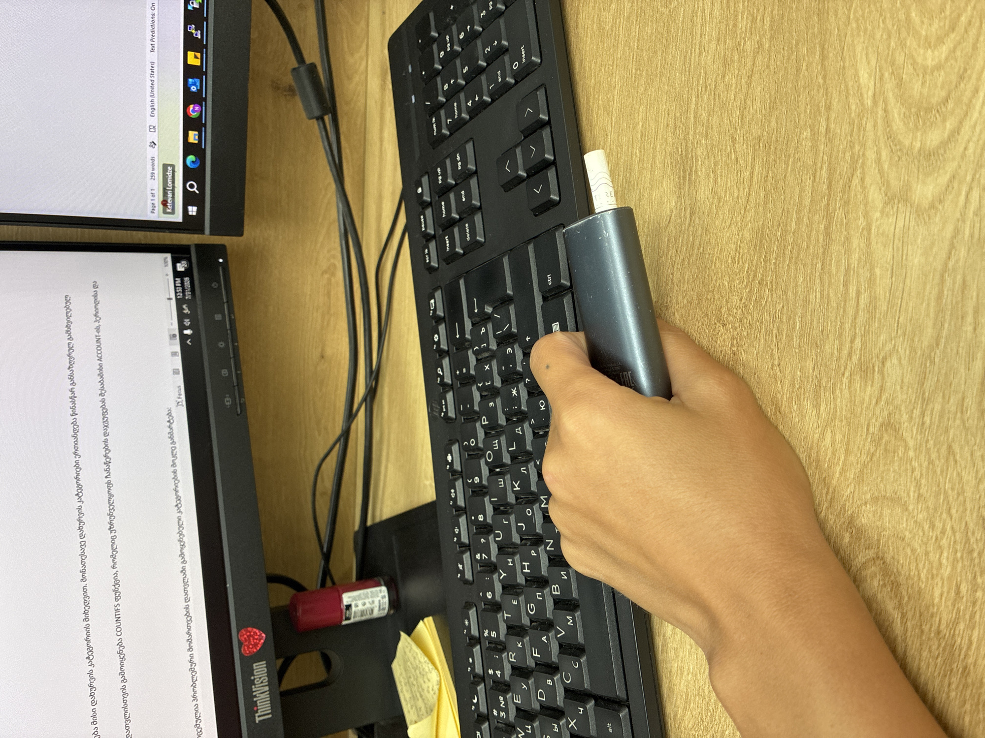 A hand is holding a USB drive over a black keyboard in an office environment. The keyboard is positioned on a wooden desk in front of two computer monitors. The left monitor displays text in a foreign language, likely Georgian, and has a "ThinkVision" logo. A red nail polish bottle is visible on the left monitor's stand. The right monitor displays a word processing document with the name "Ketevan Lomidze" and a word count. The time on the system tray of the left monitor reads "12:53 PM 7/31/2026". The scene suggests a professional setting, possibly an office in Tbilisi, Georgia, based on the presence of the Georgian language on the screen.