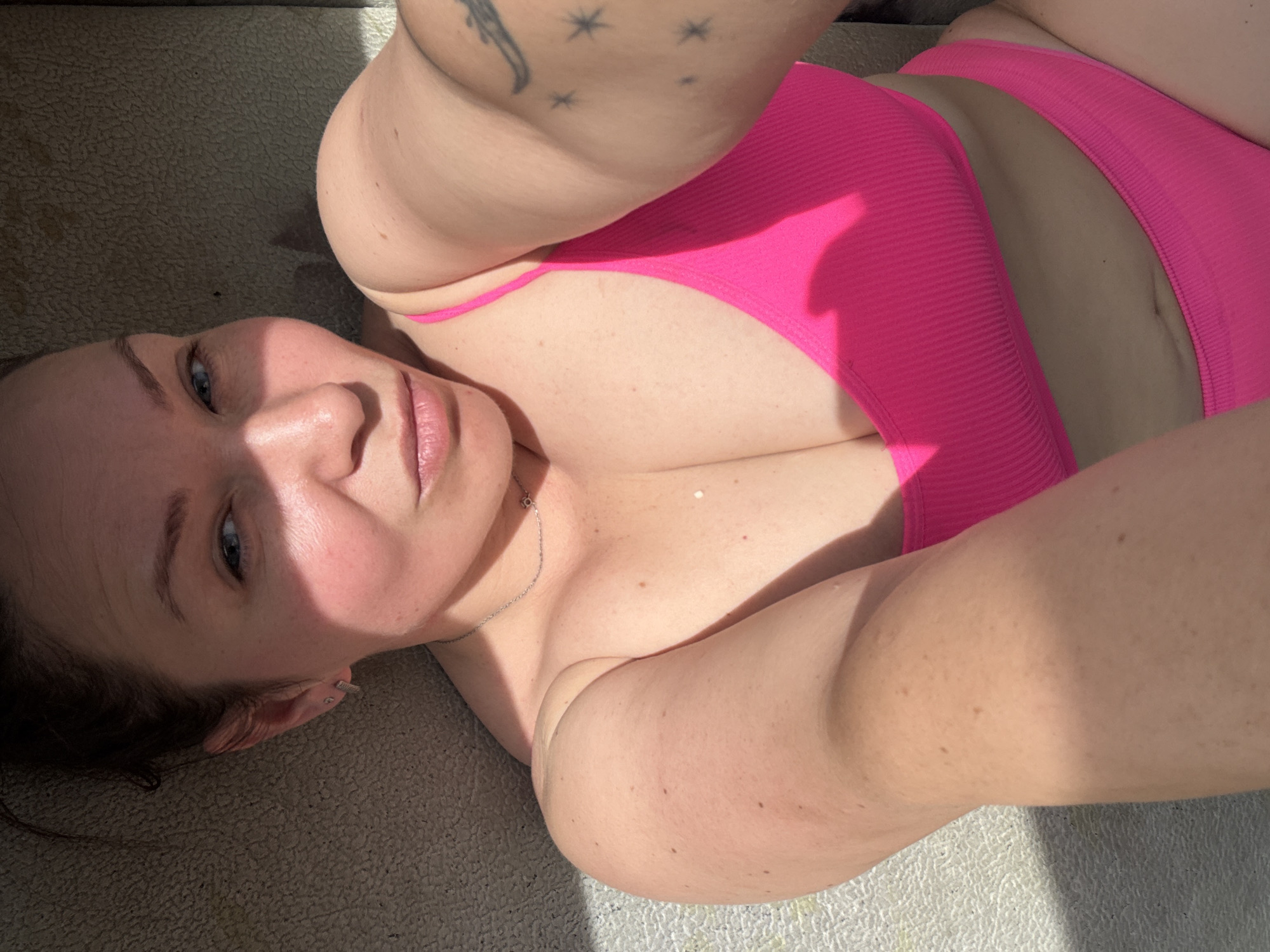 A woman lies on a textured surface, possibly fabric, wearing a pink bikini. She is fair-skinned with blue eyes and brown hair pulled back. A silver necklace is visible on her chest. A tattoo of stars and another design is on her arm. Sunlight illuminates the scene, creating shadows on her face and body. The location in Chaikovsky, Russia is not discernible from the image.