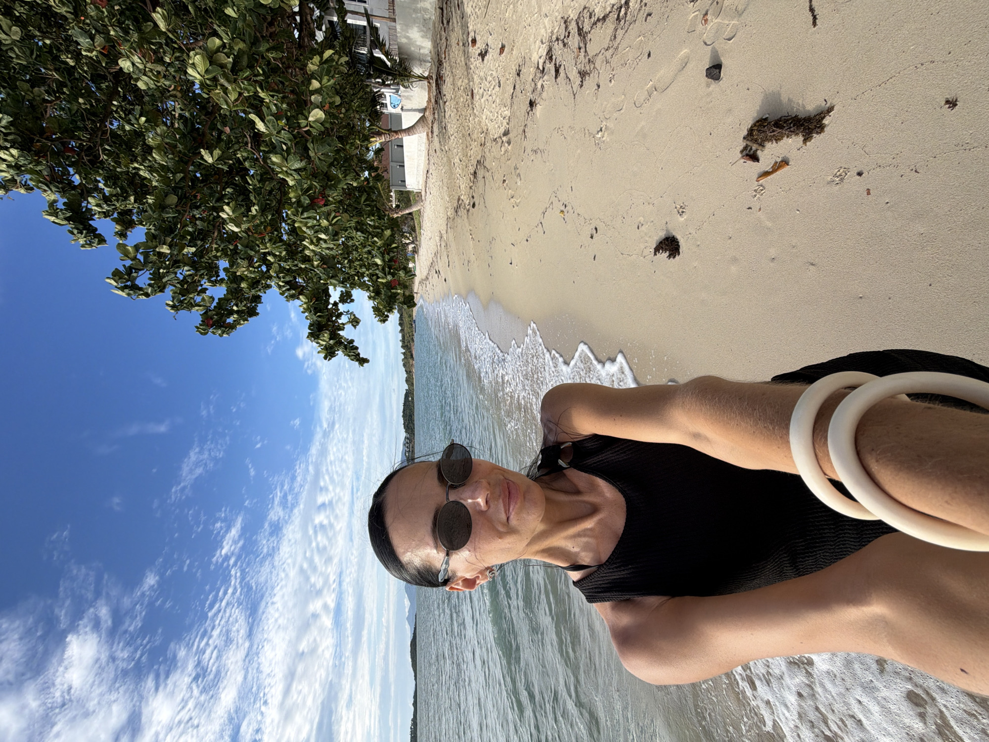 A woman with dark hair pulled back, wearing sunglasses and a black sleeveless top, stands on a sandy beach at the edge of the water, taking a selfie. The ocean is to her left, with gentle waves lapping the shore. To her right is a stretch of sand with some seaweed and debris, leading to a line of palm trees and buildings. The sky above is blue with scattered white clouds. The location is Ko Samui District, Thailand.