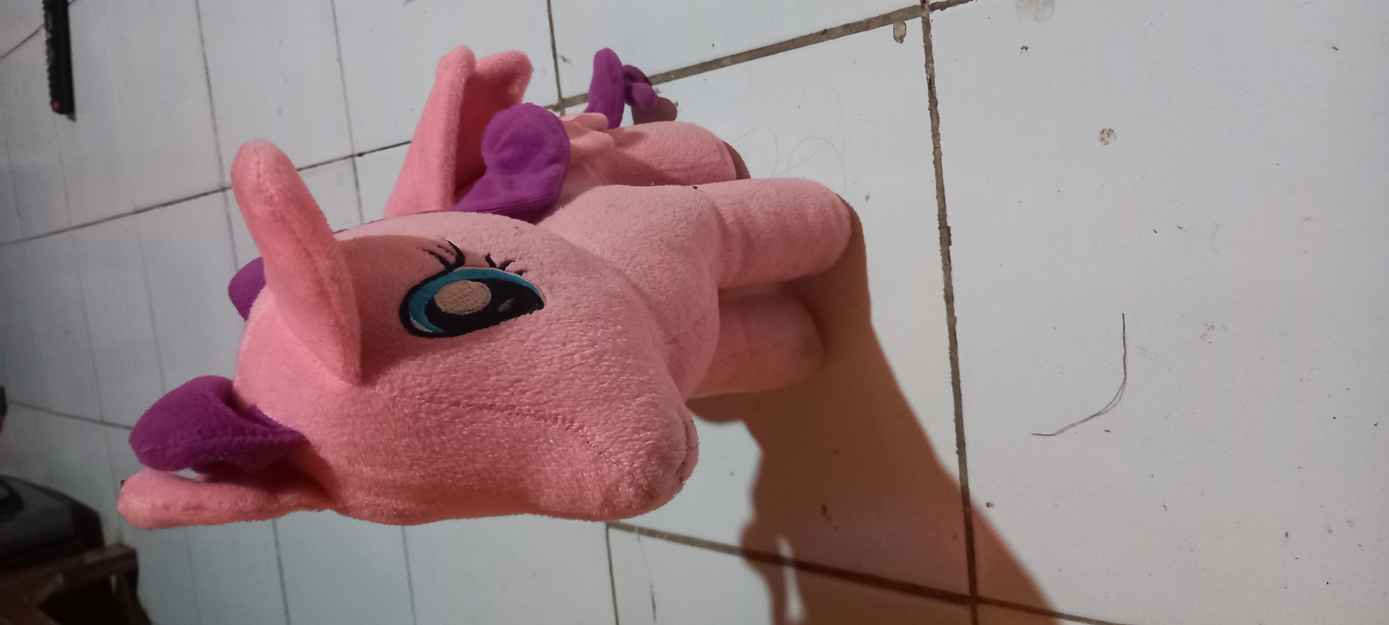 A close-up shot focuses on a pink plush toy unicorn with purple accents, positioned on a tiled floor. The toy is oriented to the left, with its head slightly lowered. Its eyes are large and blue with black outlines and eyelashes. The unicorn has two pointed ears and two small, wing-like structures on its back, all in purple and pink. The floor is made of white square tiles, some of which show signs of wear and dirt. A faint shadow of the toy is cast to the left. The background is blurred, but a dark object and part of a wooden structure are visible, suggesting an indoor setting. The overall scene depicts a simple domestic environment, likely in Gulurejo, Indonesia, given the context provided.