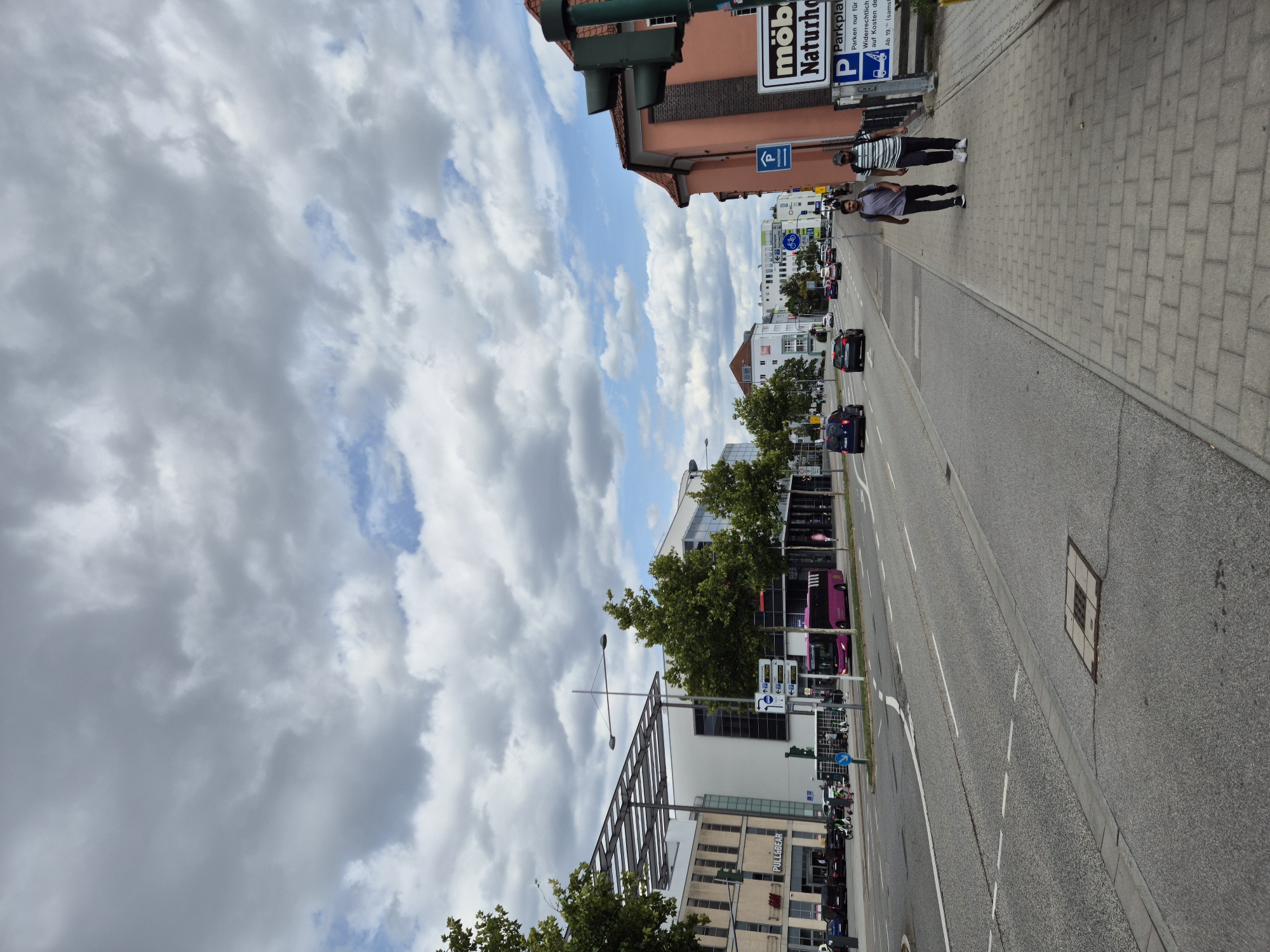 The image captures a street scene in Regensburg, Germany, under a cloudy sky with patches of blue. On the left side of the street, modern buildings with glass facades and prominent signage, including "PULL&BEAR," stand adjacent to a multi-story building. Across the street, a pink bus is visible, along with cars and bicycles. The right side of the street features a sidewalk with two men walking. A traffic light is prominently displayed near a sign for "Parkplatz" (parking lot) and "möbel naturhof" (furniture naturhof). Buildings with traditional architecture, including a pink-hued structure, are also present. The road is paved with clear lane markings and a drain cover.