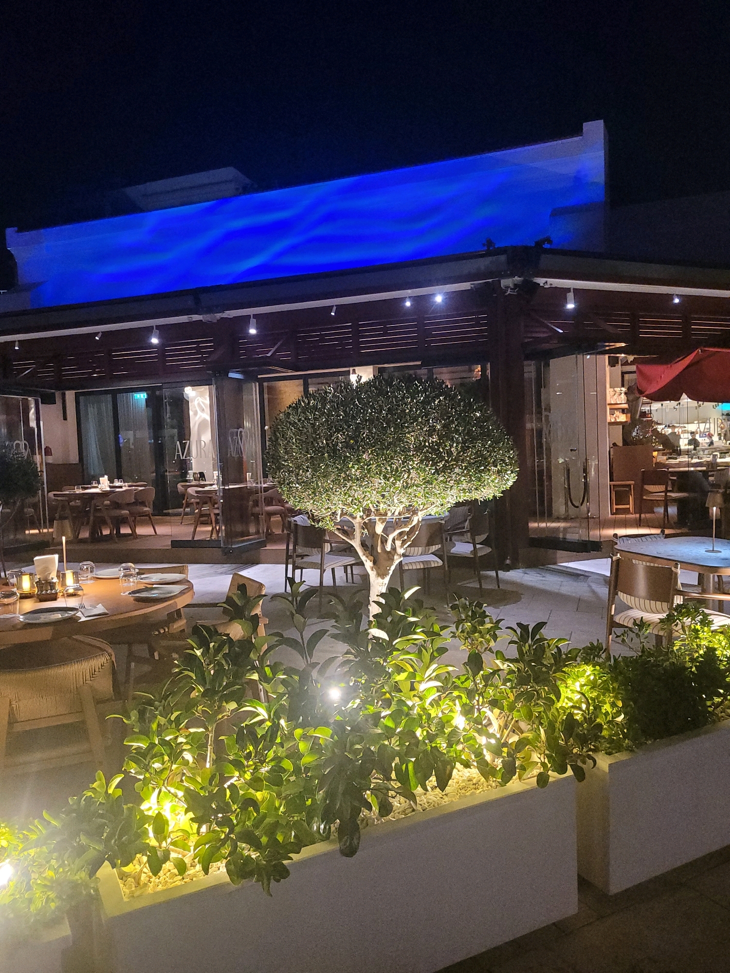 The image depicts the exterior of a restaurant named "AZURA" in Limassol, Cyprus, at night. The restaurant has an outdoor seating area with tables and chairs. The architecture features a dark wooden roof structure with modern lighting. A prominent blue illuminated panel is visible on the building's facade, creating a vibrant, atmospheric effect. In the foreground, well-lit greenery is planted in white rectangular planters. Several tables are set for dining, with plates, cutlery, and glasses. The overall ambiance suggests a sophisticated dining establishment with an emphasis on outdoor ambiance.
