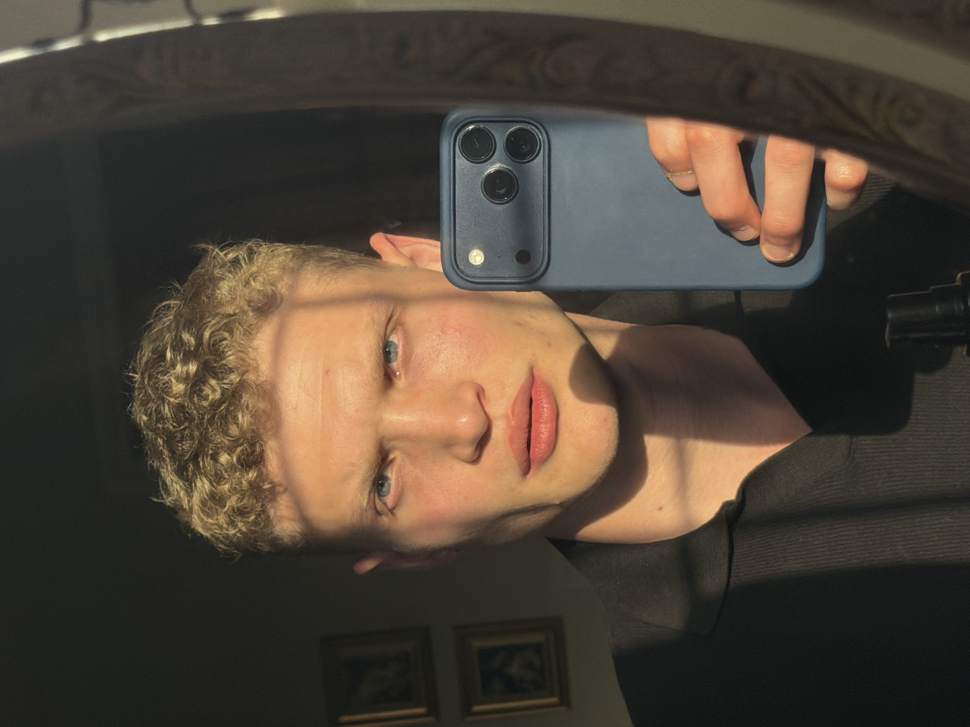 A person is taking a selfie in a mirror. The person is fair-skinned with blonde curly hair and blue eyes. They are holding a blue smartphone with three camera lenses and a flash in their right hand, partially obscuring their face. They are wearing a dark-colored garment. Two framed pictures are visible on the wall behind them. The environment appears to be indoors, lit by a strong light source that creates stark shadows. There is no indication of the location being in East Suffolk, United Kingdom, or any other location.