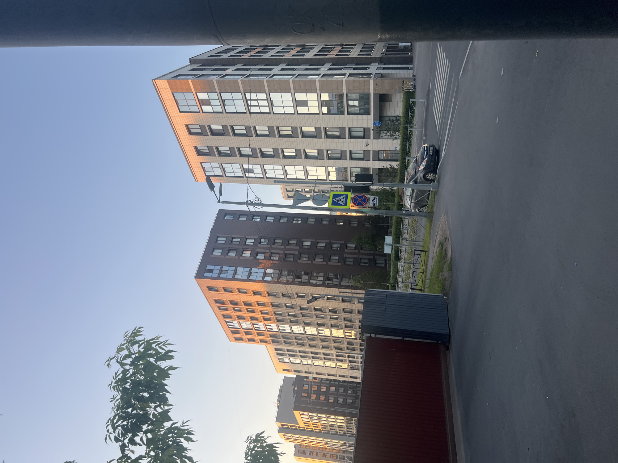The image depicts a modern urban street scene in the city of Unknown, Unknown, captured during what appears to be late afternoon or early evening, evidenced by the warm, low-angle sunlight illuminating building facades.
In the mid-ground, a series of multi-story contemporary buildings line the street. The two prominent buildings to the right feature light beige and darker grey paneling, with numerous large, reflective windows that prominently reflect the golden sky and sunlight. The building on the far right is partially obscured by a dark, large cylindrical object, likely a pillar or utility pole, which dominates the extreme right edge of the frame. Further to the left and in the background, several more buildings exhibit dark brown or reddish-brown facades, with many windows also reflecting the warm light.
The foreground consists of a dark asphalt road with a marked pedestrian crosswalk. A dark-colored sedan is visible parked or moving slowly near the crosswalk on the left side of the road. On the sidewalk to the right, several yellow and black electric scooters are parked.
Street furniture includes a tall lamppost with a dark fixture, positioned between the central buildings. Beneath the lamppost, a pole holds a set of traffic signs, including a pedestrian crossing sign (blue square with white figure) and a "No Stopping" sign (red circle with diagonal line). Metal barrier fences run alongside the sidewalk and in the distant background.
To the far left in the foreground, a dark red metal fence or gate is partially open, revealing a smaller, dark grey corrugated structure behind it. Green tree branches with leaves extend into the upper left portion of the frame. Small hedges and patches of grass are visible along the base of the buildings and sidewalks. The sky overhead is clear, displaying a light blue hue that transitions to a warmer, lighter tone near the horizon.