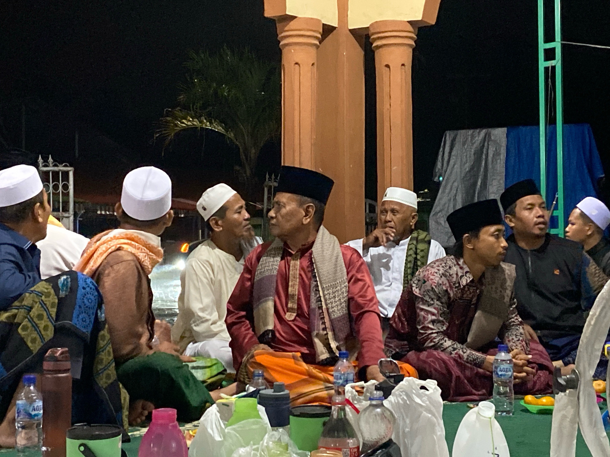 The image depicts a nighttime gathering of several Muslim men, likely in Pantai Gading, Indonesia, seated on the ground. The scene appears to be near a building with large, round, brown pillars, possibly a mosque or a religious institution. The men are wearing traditional Islamic attire, including peci (skullcaps) and some are adorned with scarves or sarongs.
In the foreground, several individuals are visible. One man in a maroon shirt and orange sarong is the central figure, wearing a patterned scarf. To his left, a man in a brown sweater and a light orange scarf is seated. In front of them are various objects, including water bottles, green and black containers, and a pink bottle. Plastic bags are also scattered on the ground, containing what seem to be food items or supplies.
Further back, other men are seen wearing white peci and traditional clothing. Some are engaged in conversation, while others appear to be listening or observing. The lighting suggests it is nighttime, with artificial lights illuminating the scene. In the background, a large white tent or tarp is visible, along with some greenery from trees. A green metal structure is also present on the right side of the image. The overall atmosphere suggests a communal gathering, possibly for religious or social purposes.