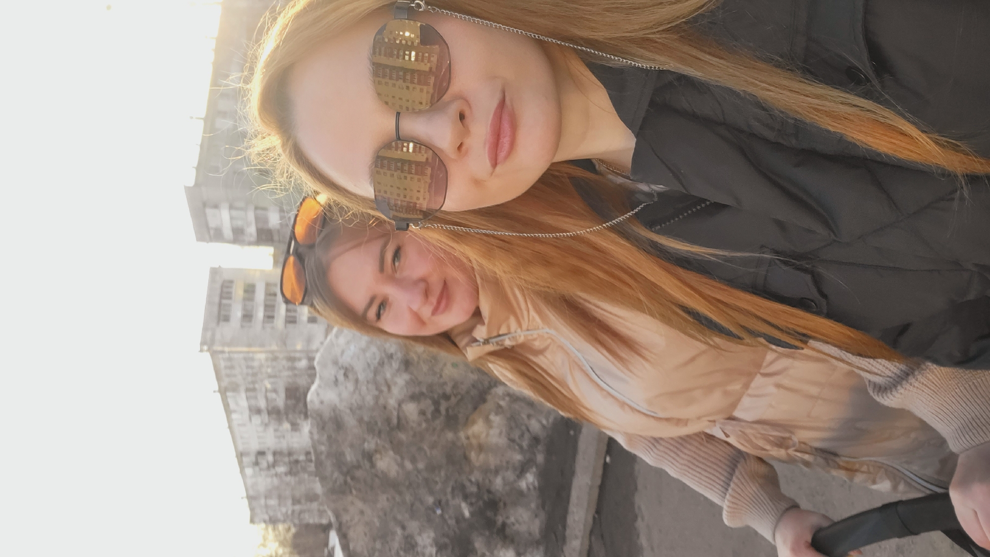 Two women with long, light-colored hair are posing for a selfie. The woman in the foreground wears dark-rimmed sunglasses with buildings reflected in the lenses and a black vest. Behind her, the second woman wears orange-rimmed sunglasses and a beige vest. Buildings are visible in the background. The location appears to be Chaikovsky, Russia.