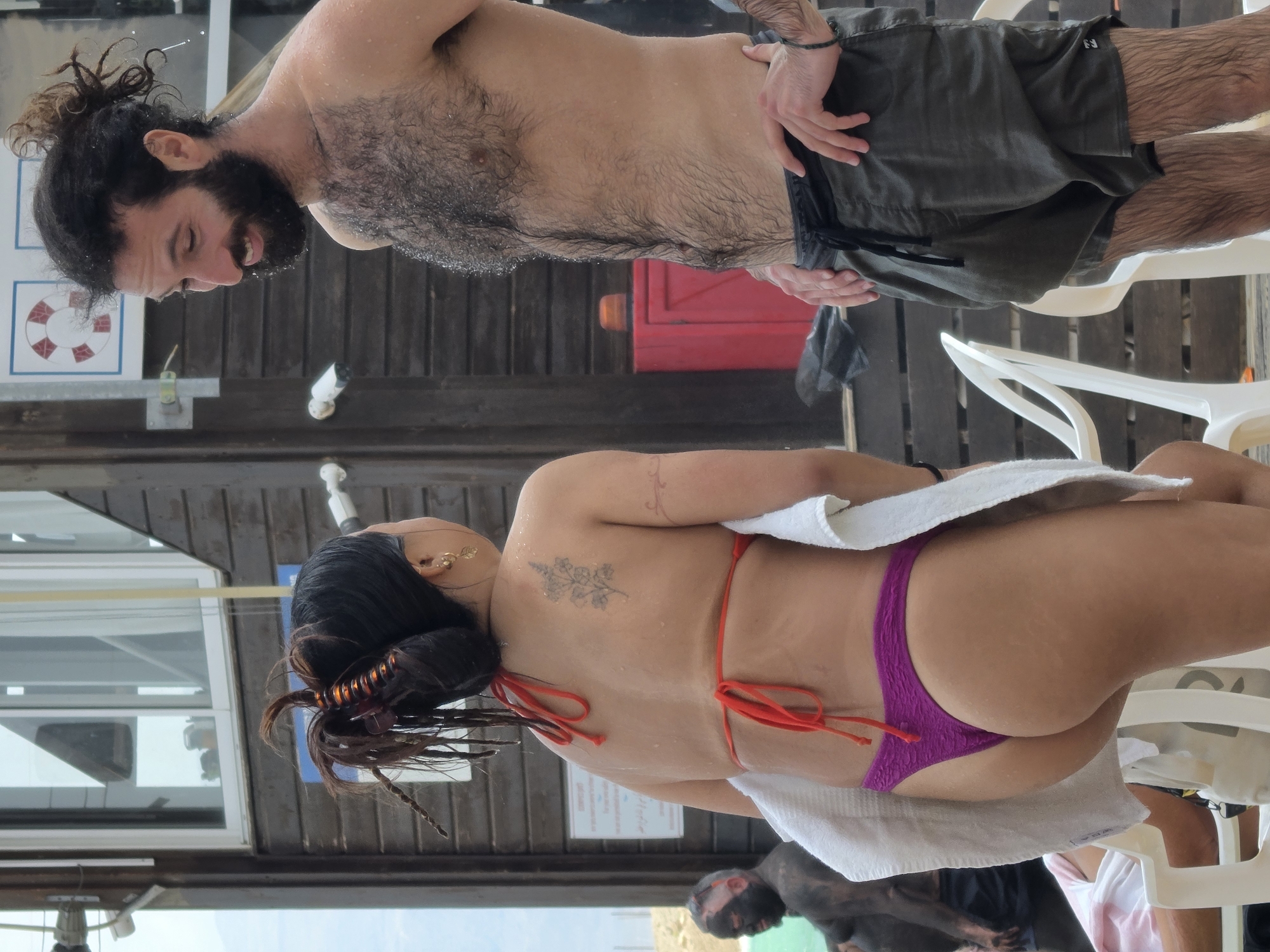 The image depicts a beach setting in Tamar Regional Council, Israel. A shirtless man with a beard and a topknot stands to the left, wearing dark-colored shorts. Adjacent to him, a woman in a purple bikini and a white towel sits on a white plastic chair. The background includes wooden paneling, a red box-like object, and glimpses of the sky and water. In the lower portion of the image, a dark-skinned individual is visible, possibly covered in mud or similar substance.